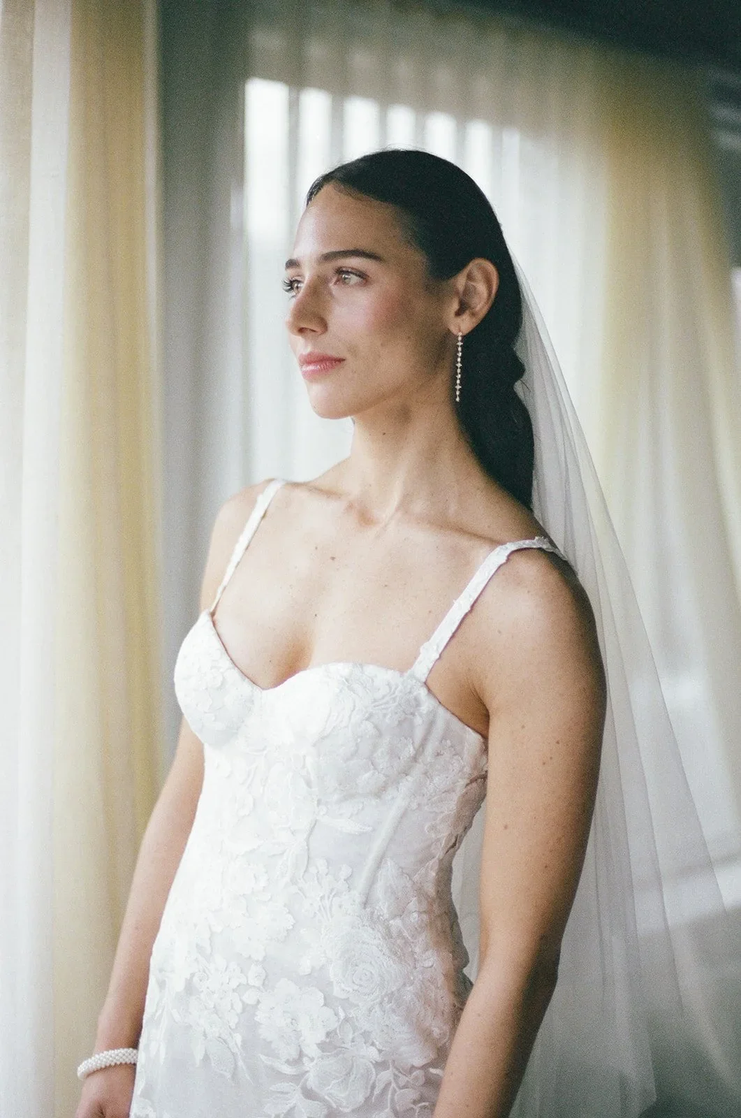 A bride with dark hair in a white lace wedding dress, looking out a window with sheer curtains, wearing a pearl bracelet and long earrings.