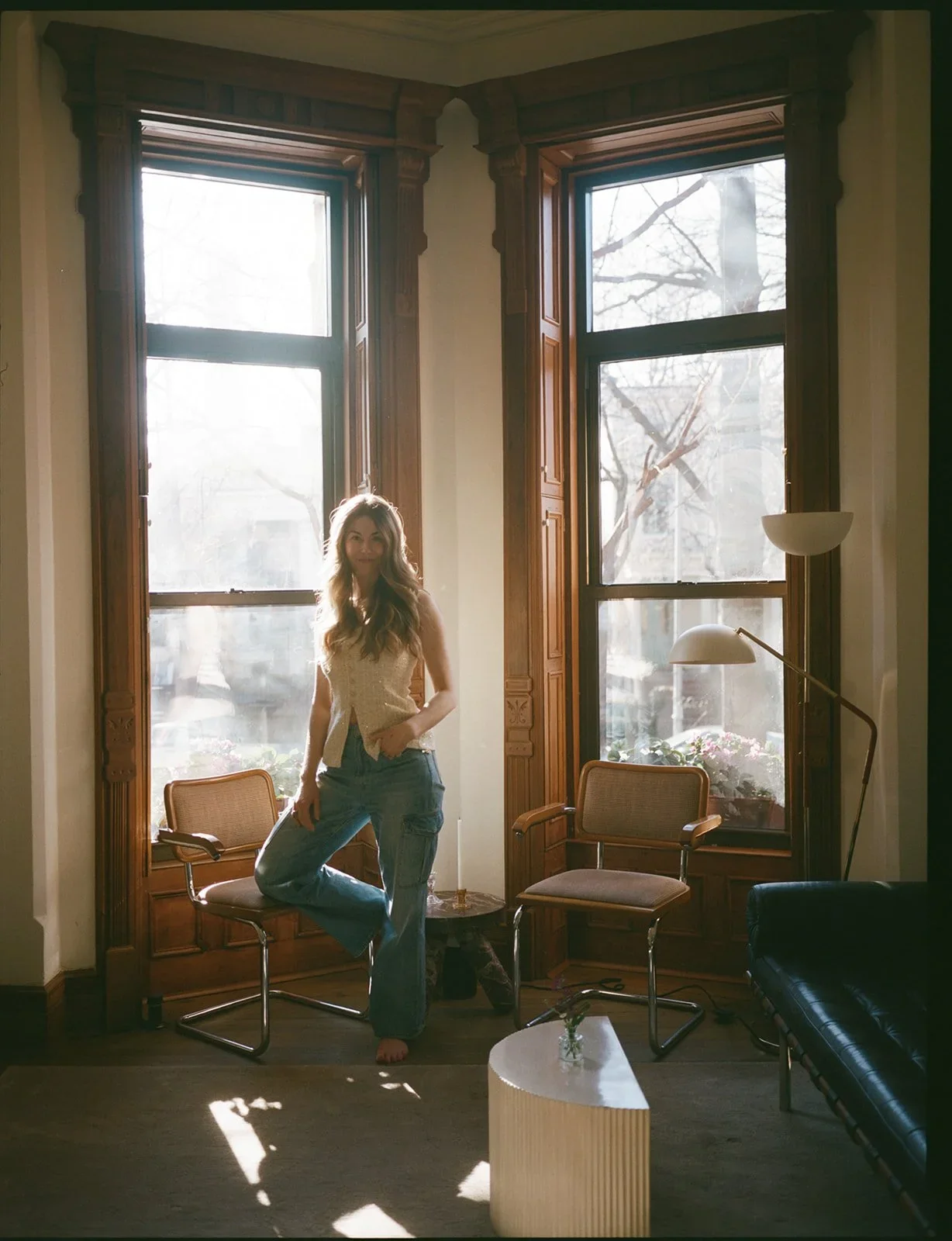 A woman sitting on a chair in a sunlit room with large windows, wooden trim, two chairs, a small table, and a black sofa.