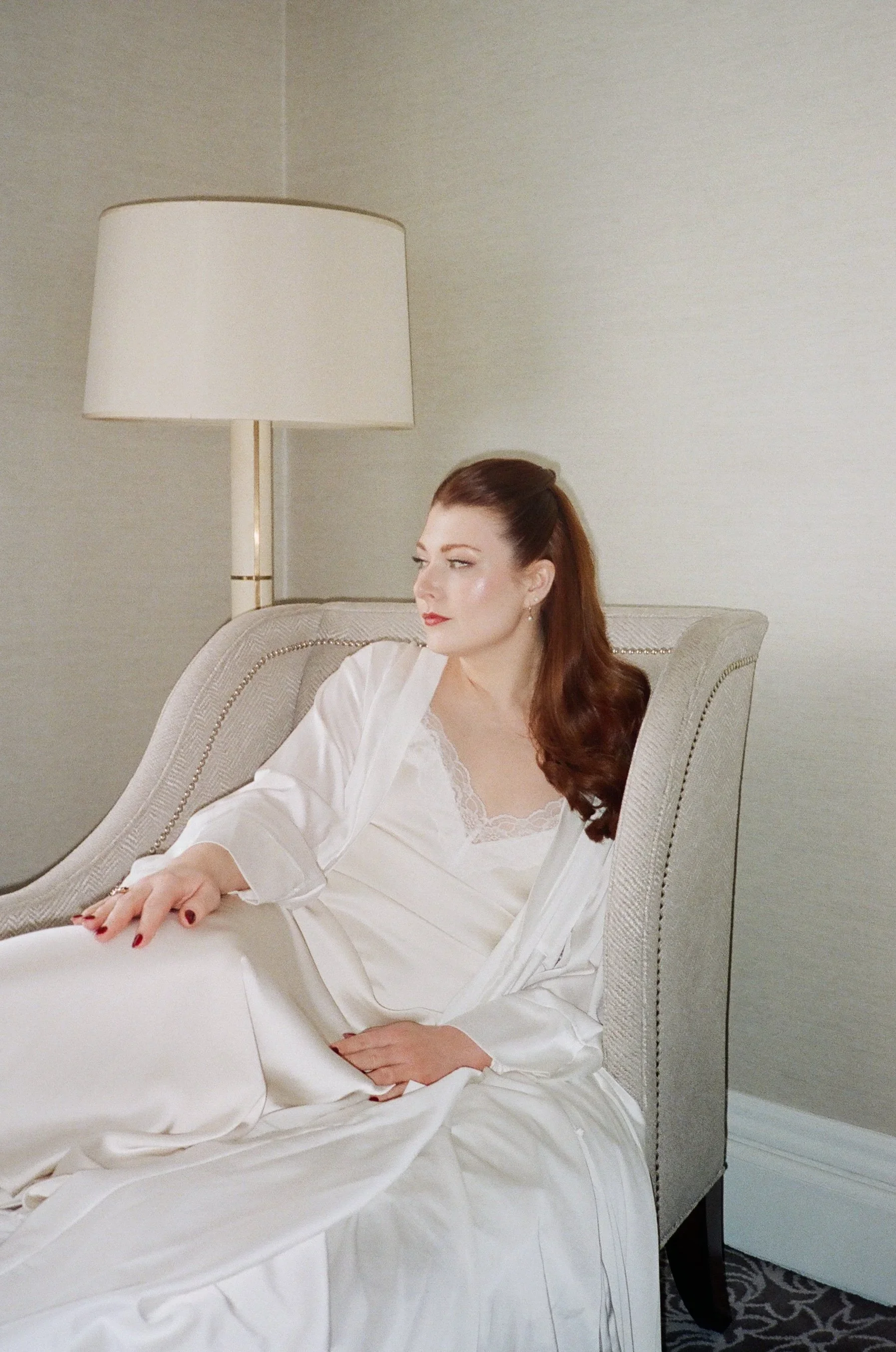 A woman with red hair wearing a white satin robe, sitting on a beige upholstered chair in a room with a lamp and light-colored wall.