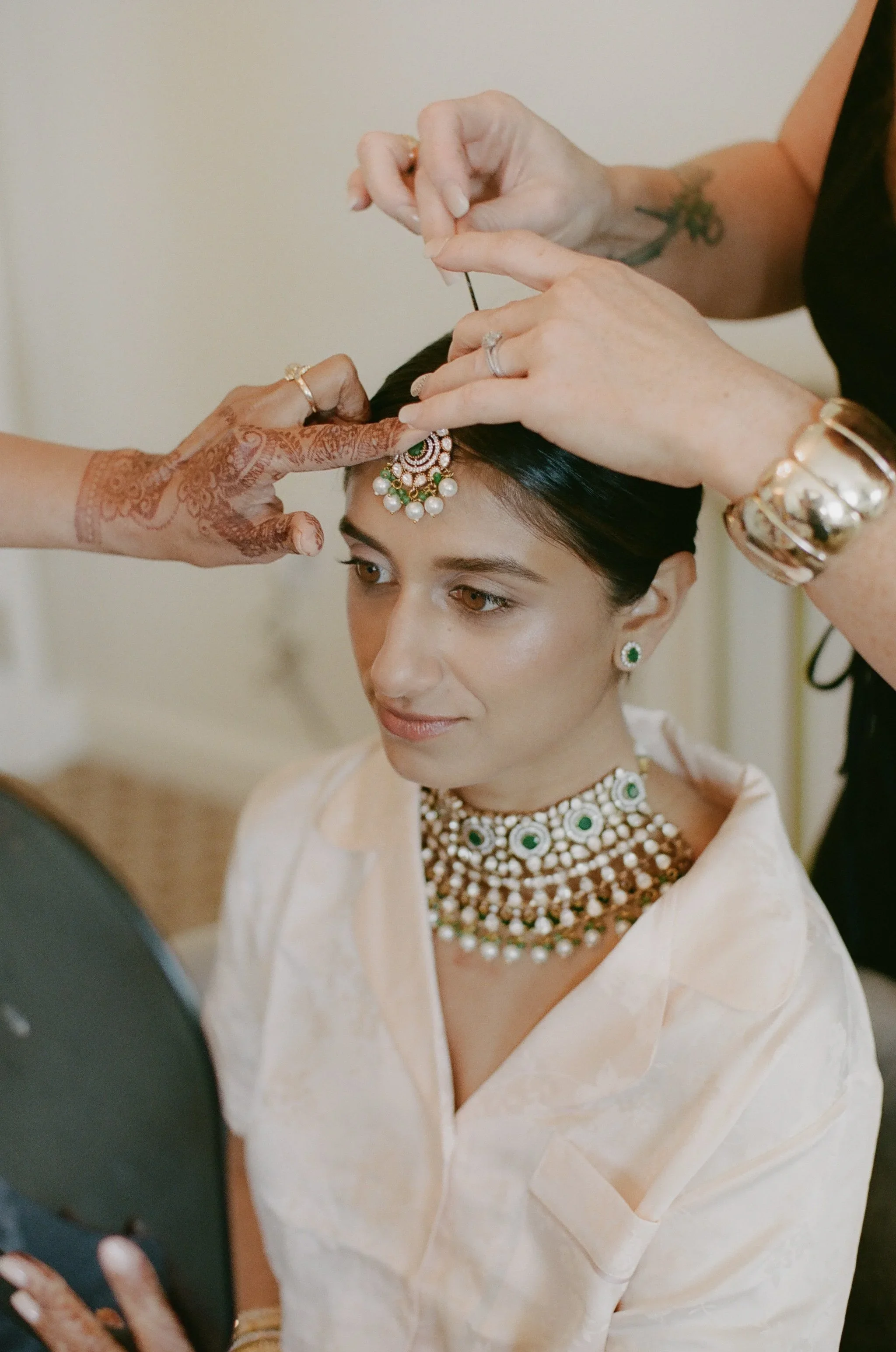 A woman getting her traditional Indian jewelry and accessories adjusted by two women, with henna on one woman's hand.