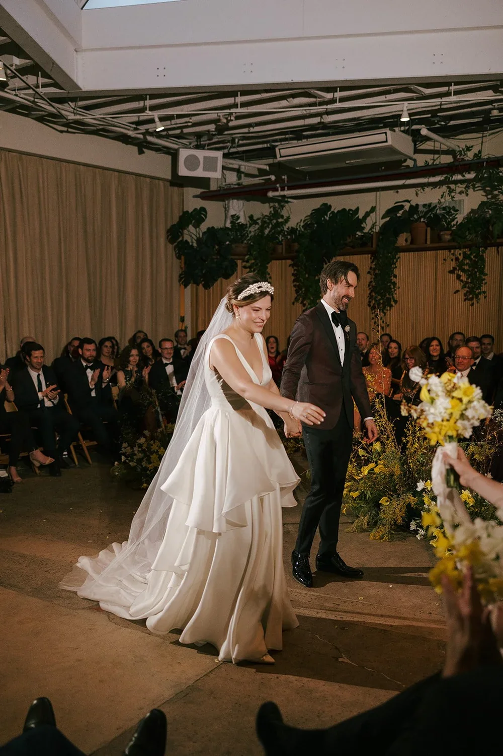 Bride and groom holding hands walking down the aisle during their wedding ceremony, surrounded by seated guests and floral decorations.