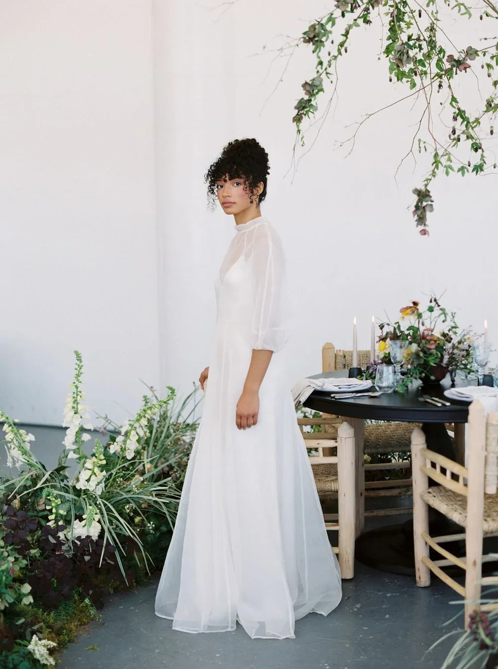 A woman in a white dress standing next to a decorated table with floral arrangements, candles, and tableware in an indoor setting with white walls and greenery.