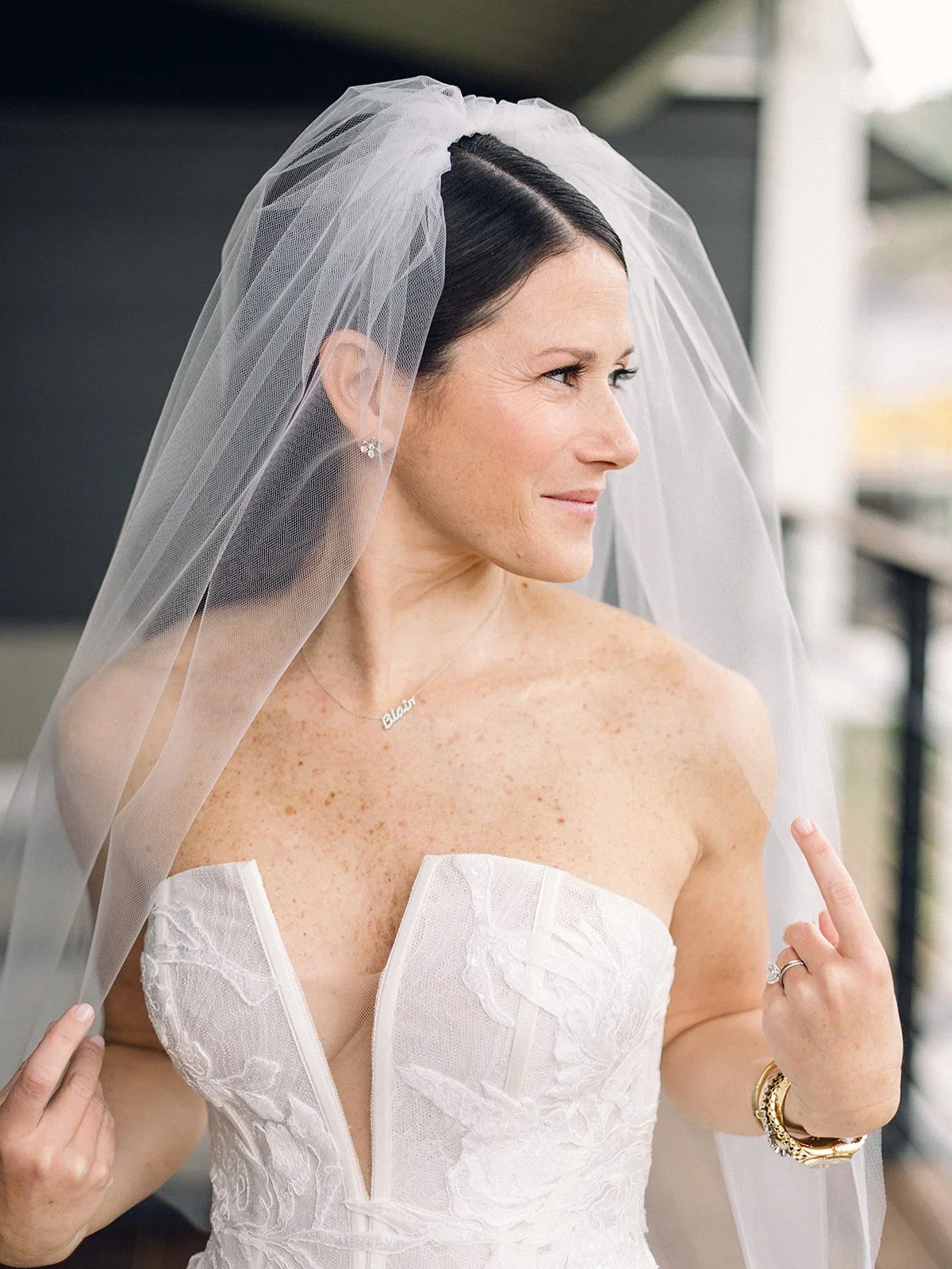 A bride with dark hair wearing a lace strapless wedding dress and a sheer veil, pointing to her jewelry.