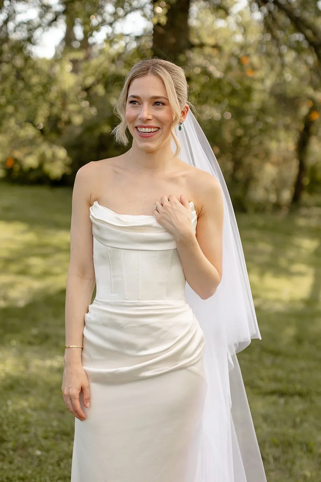 A smiling bride in a strapless white wedding dress with a long veil outdoors among trees.