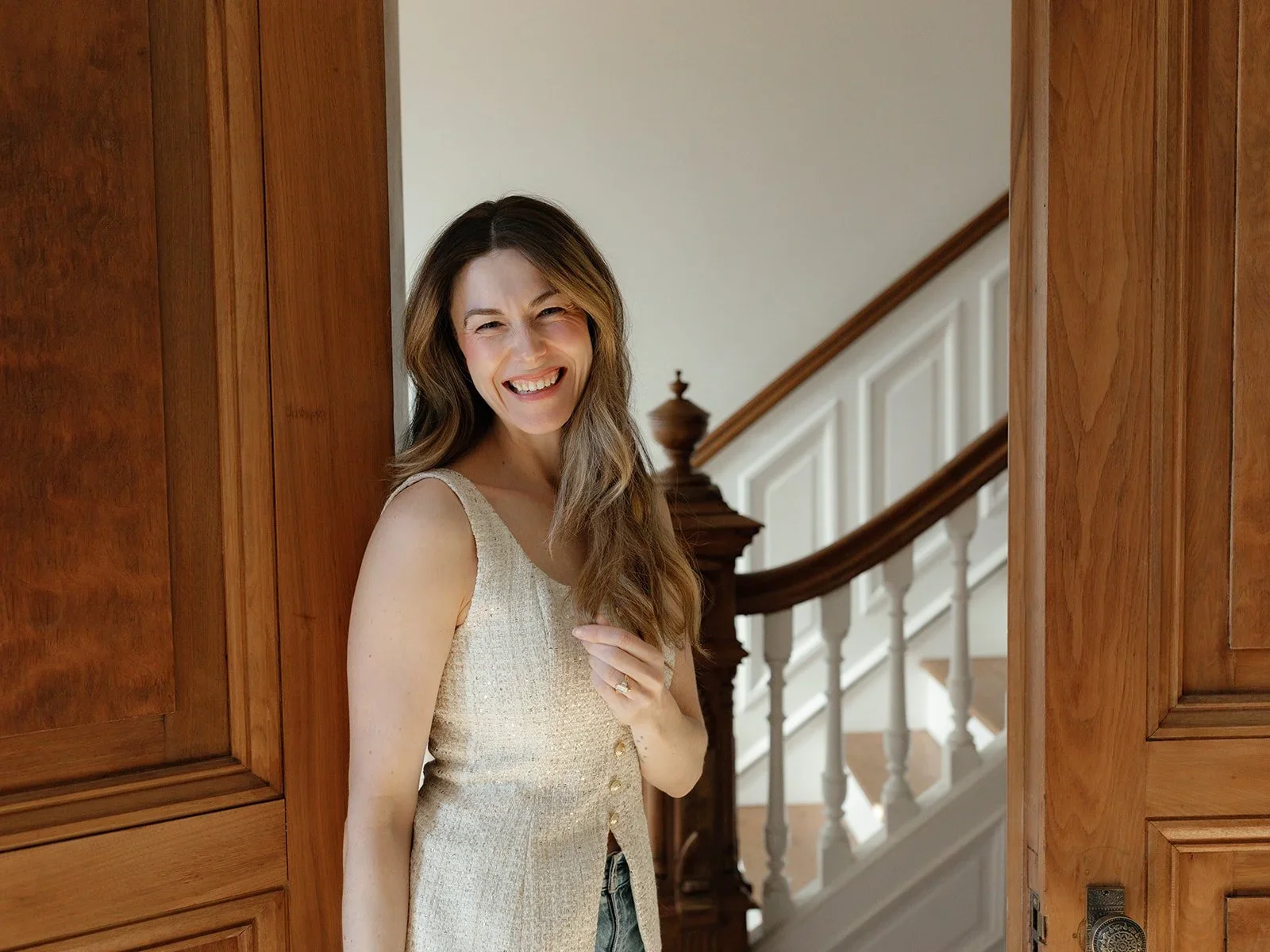 A woman with long brown hair smiling and standing in a doorway, near a staircase with a wooden handrail.