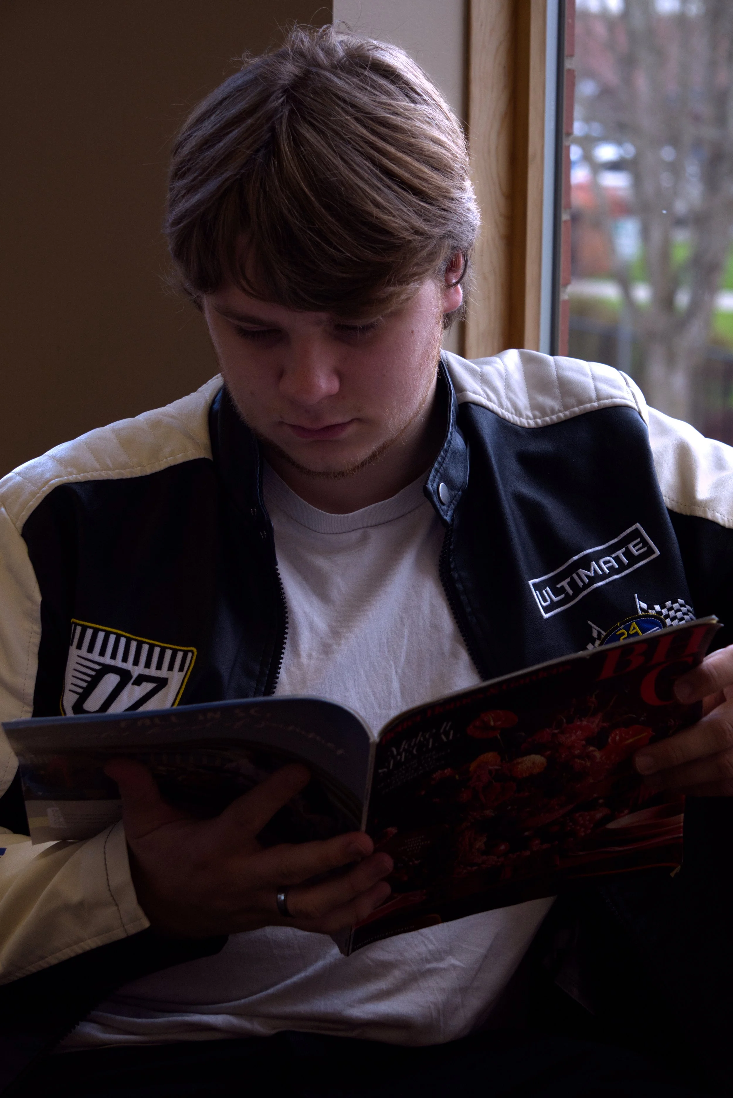 A young man with light brown hair, wearing a black and cream-colored jacket with patches, is sitting inside near a window, looking down at a magazine.