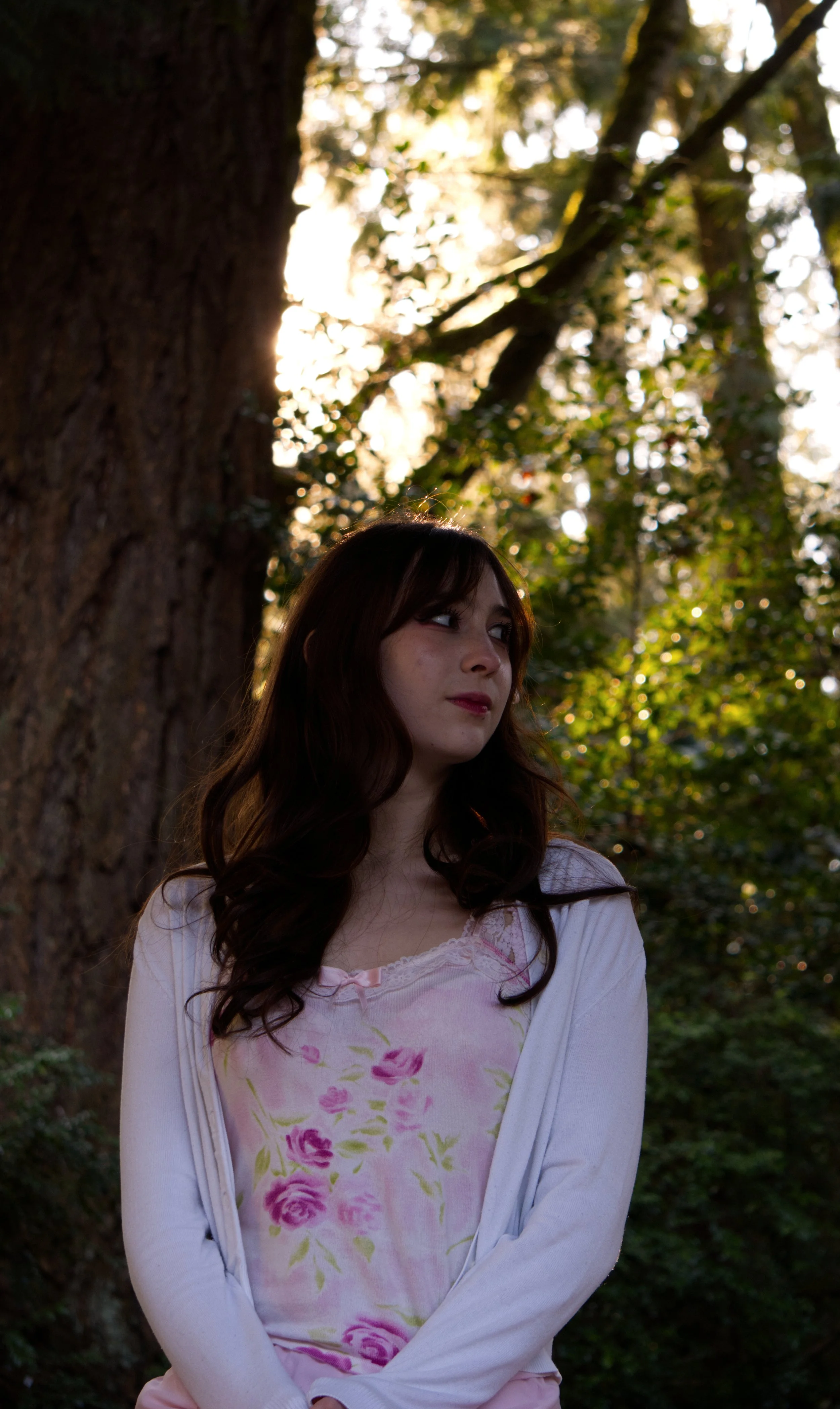A young woman with long dark hair looking to her left, standing outdoors in front of a large tree with sunlight filtering through the leaves in the background.