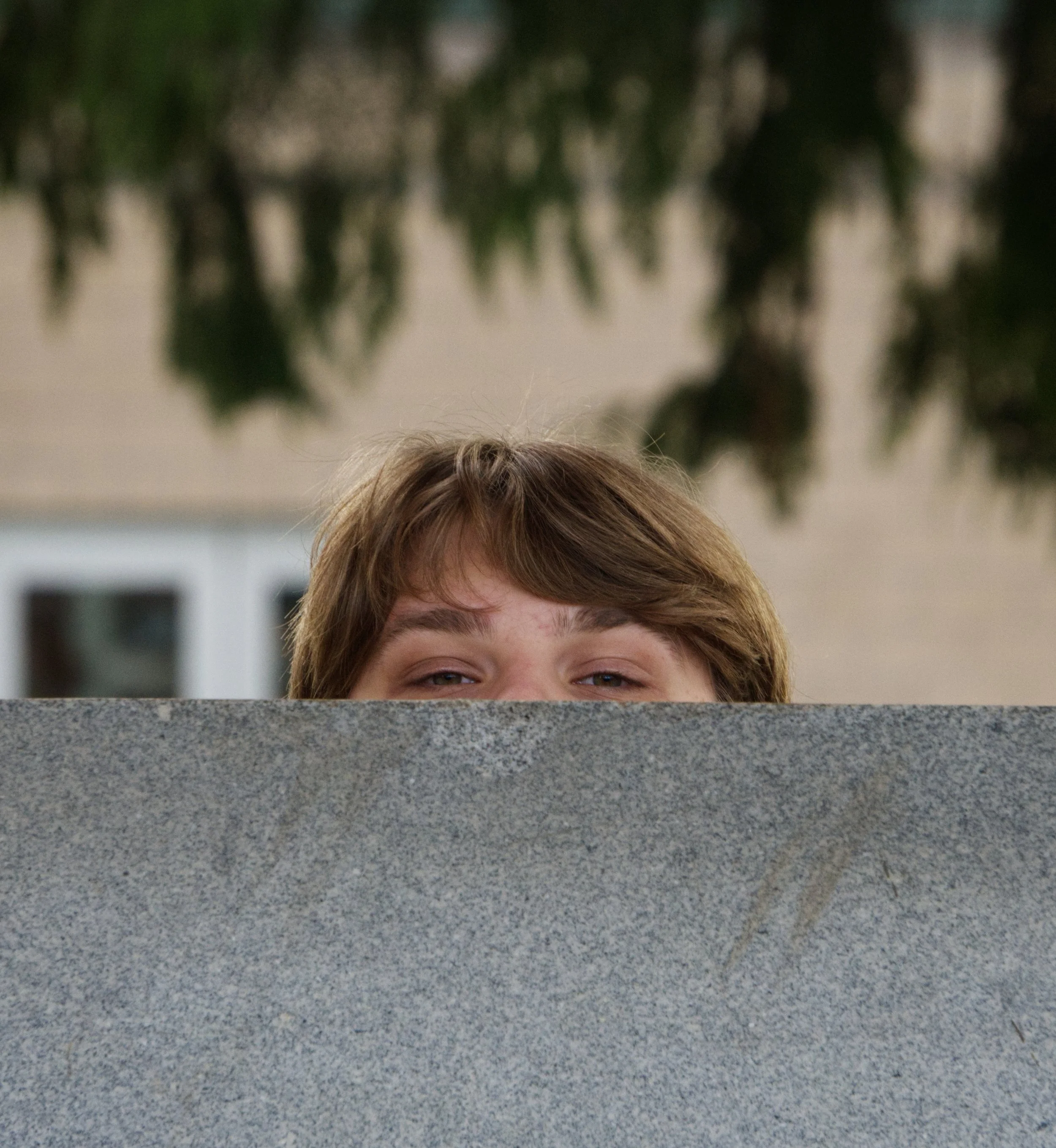 Person peeking over a gray stone wall with green foliage and a building in the background.