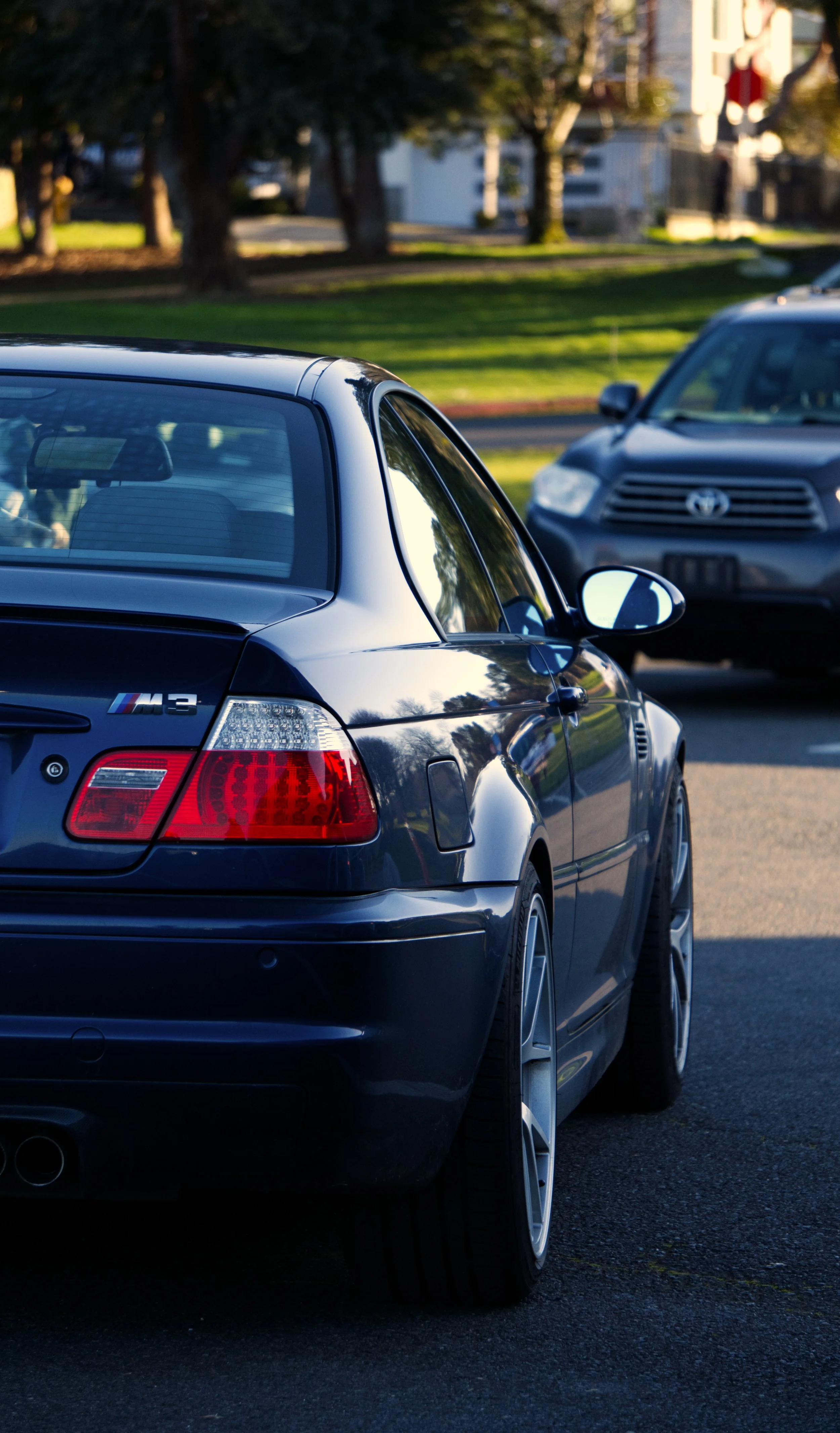 A black BMW M3 parked in a lot with a Toyota SUV parked nearby, green grass and trees in the background.