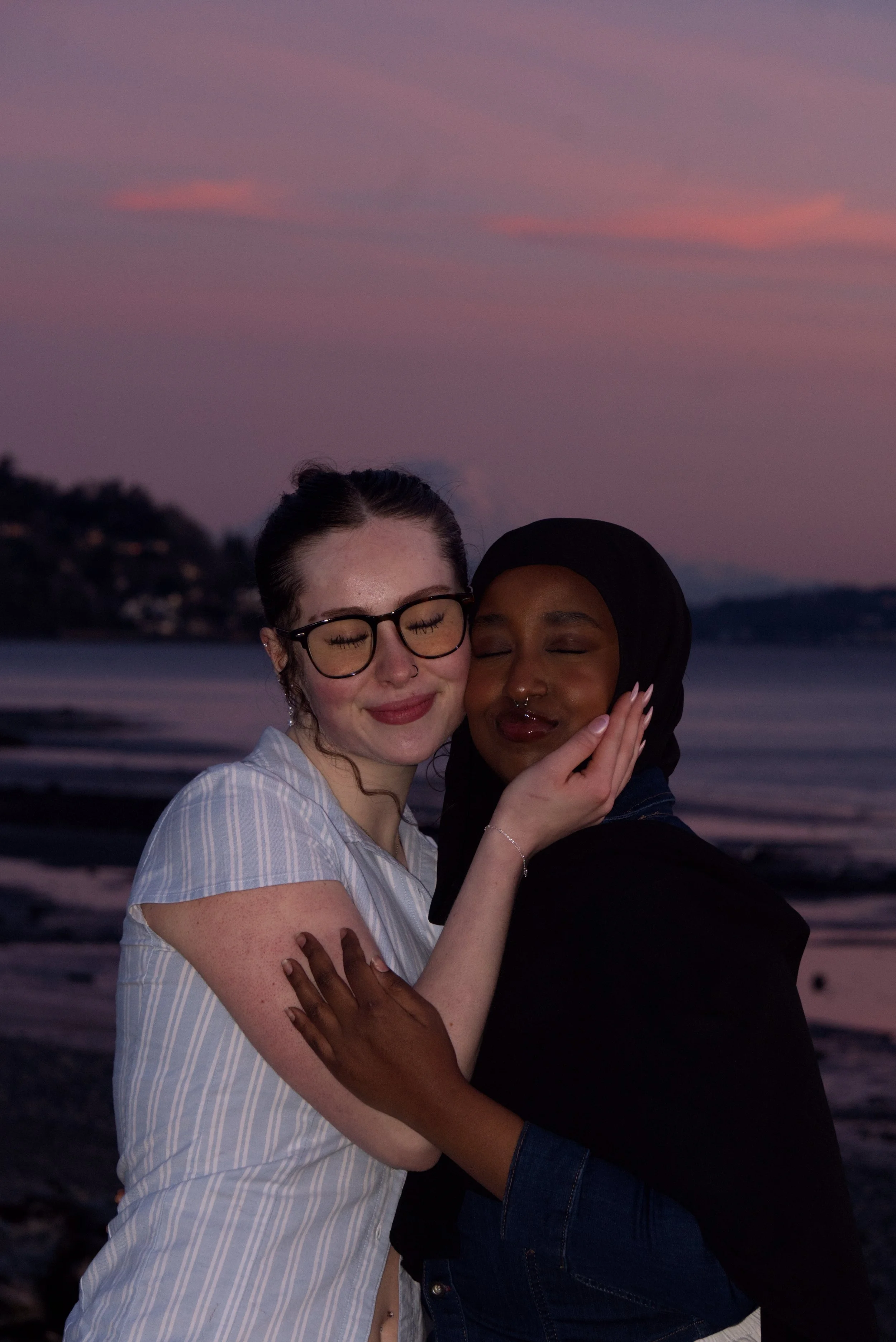Two women, one with glasses and the other with a headscarf, hugging each other with closed eyes on a beach during sunset.