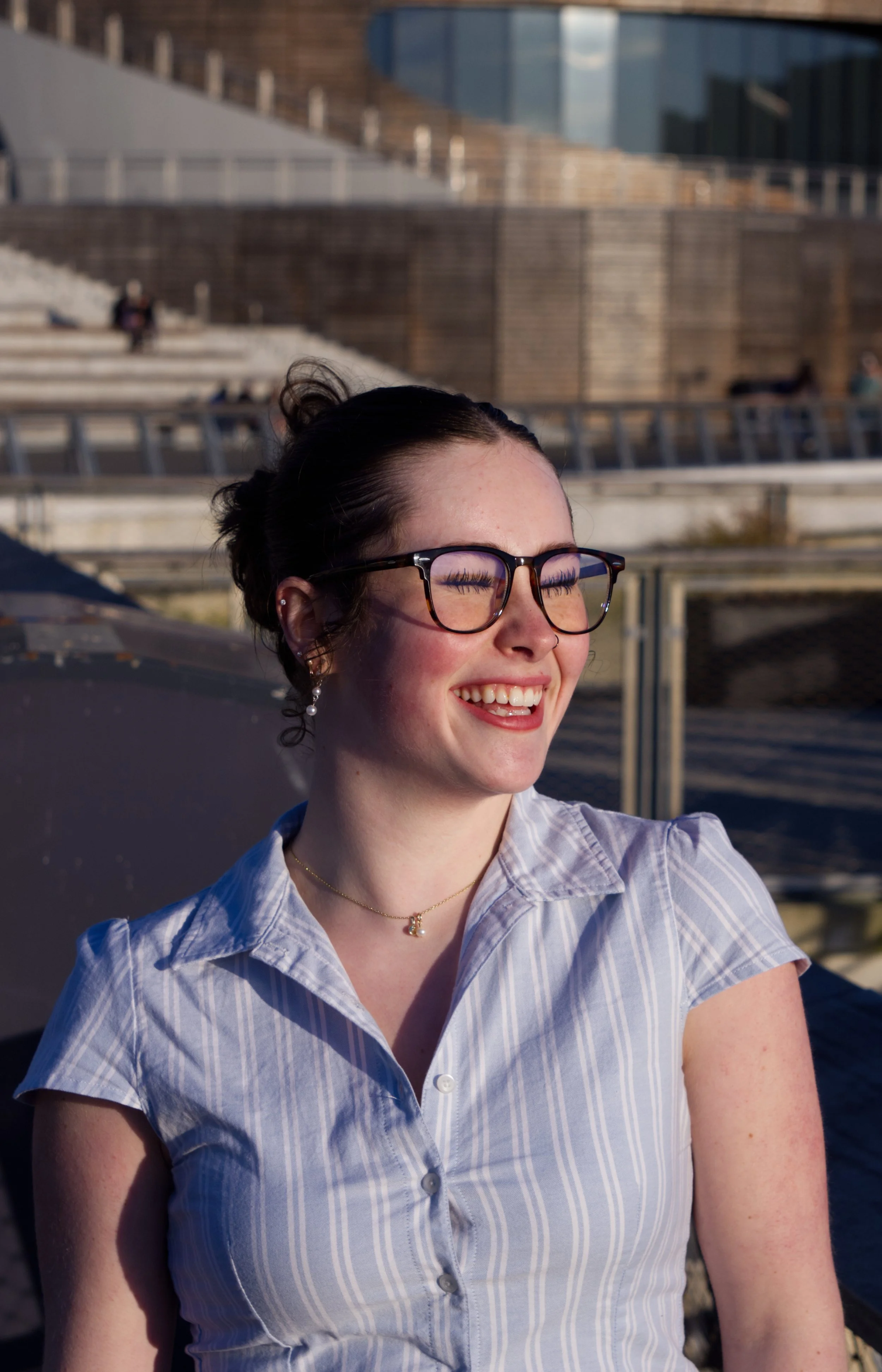A young woman with glasses smiling outdoors on a sunny day, wearing a light blue striped shirt, earrings, and a necklace, with a modern building and steps in the background.