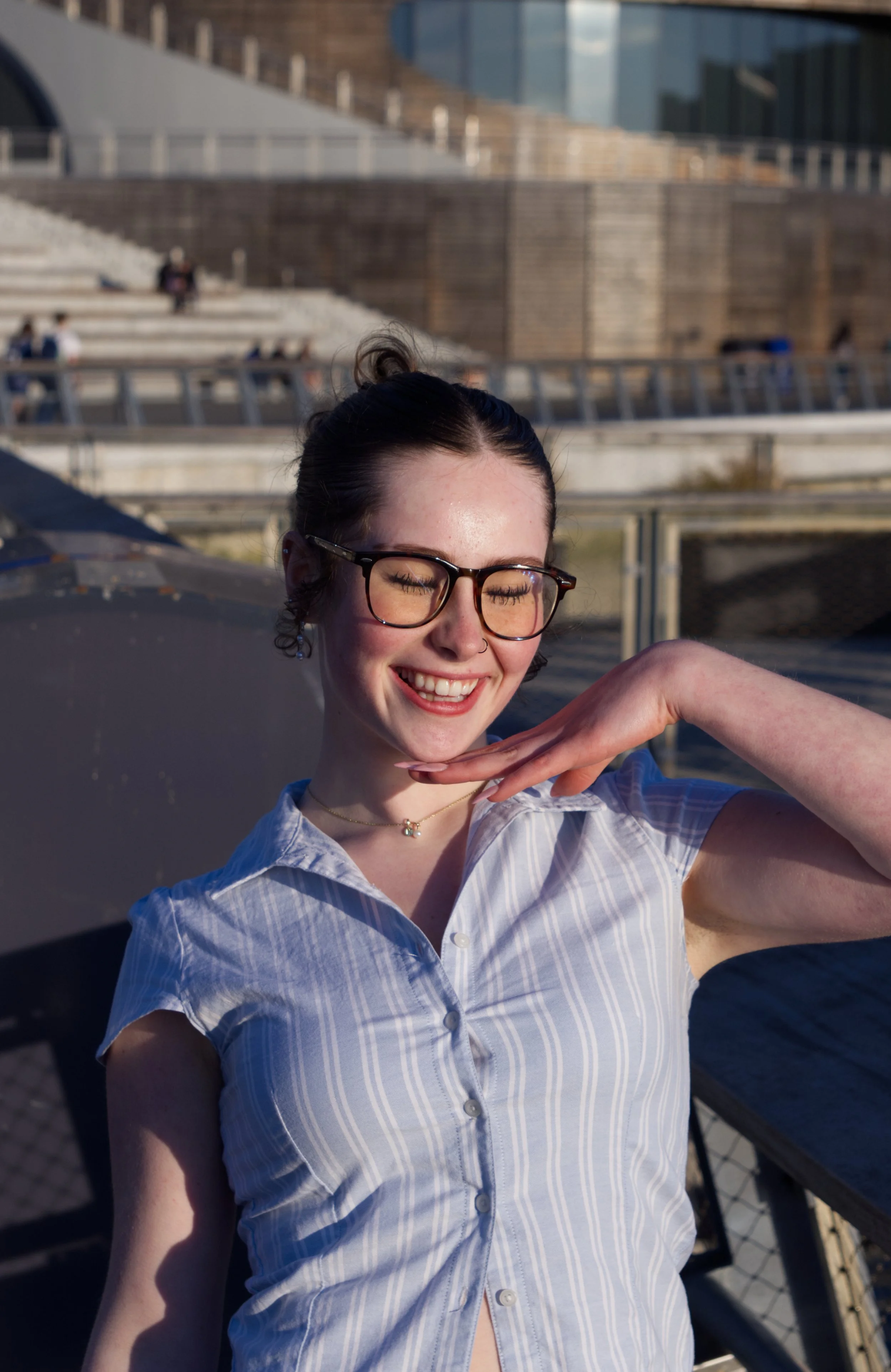 A young woman with glasses smiling and closing her eyes, posing with her hand under her chin on a rooftop with city buildings in the background.