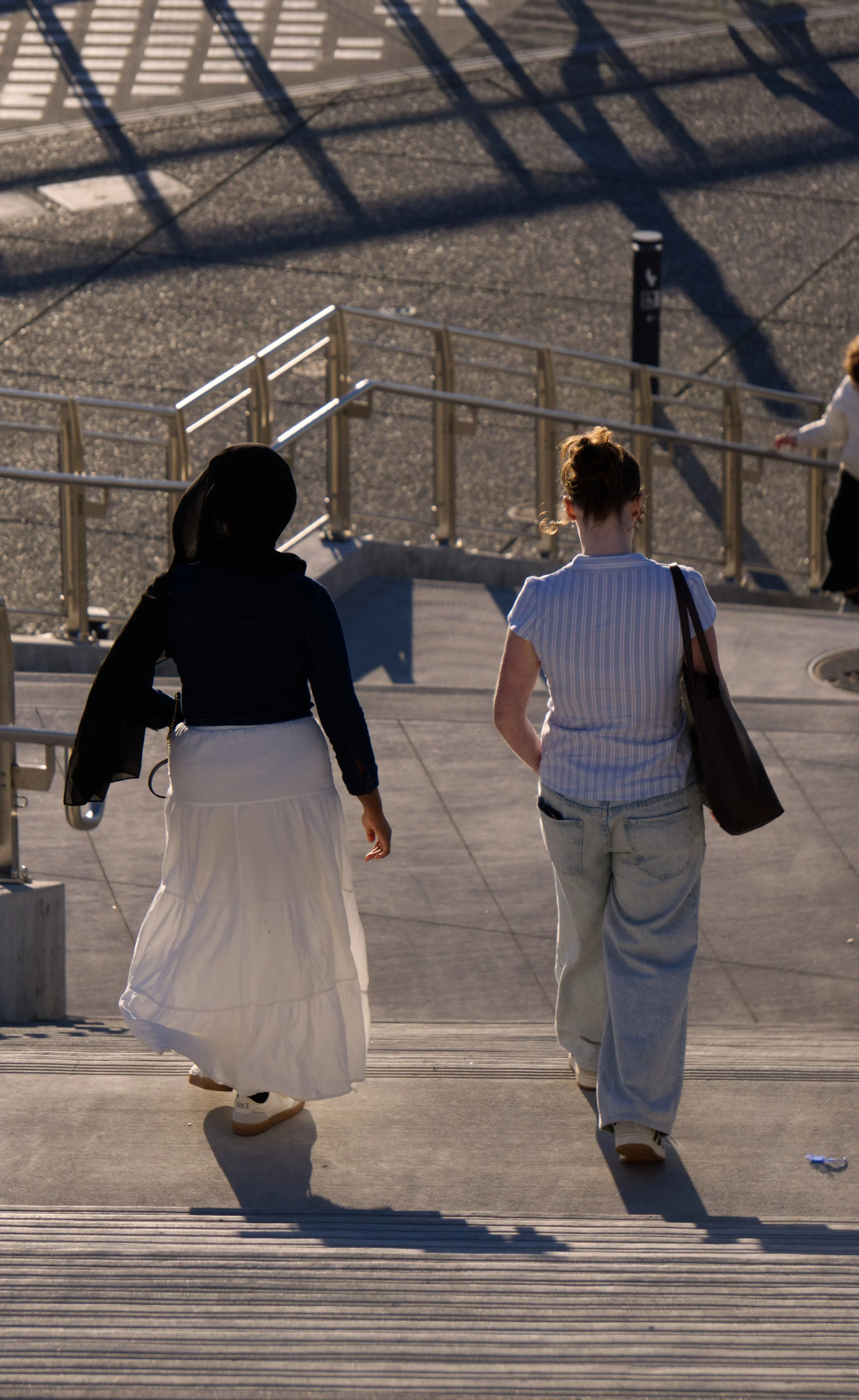 Two women walking upstairs in an outdoor urban setting during late afternoon. One is wearing a white skirt and black top with a black head covering, and the other is wearing a striped shirt with loose-fitting jeans, carrying a shoulder bag. Shadows a