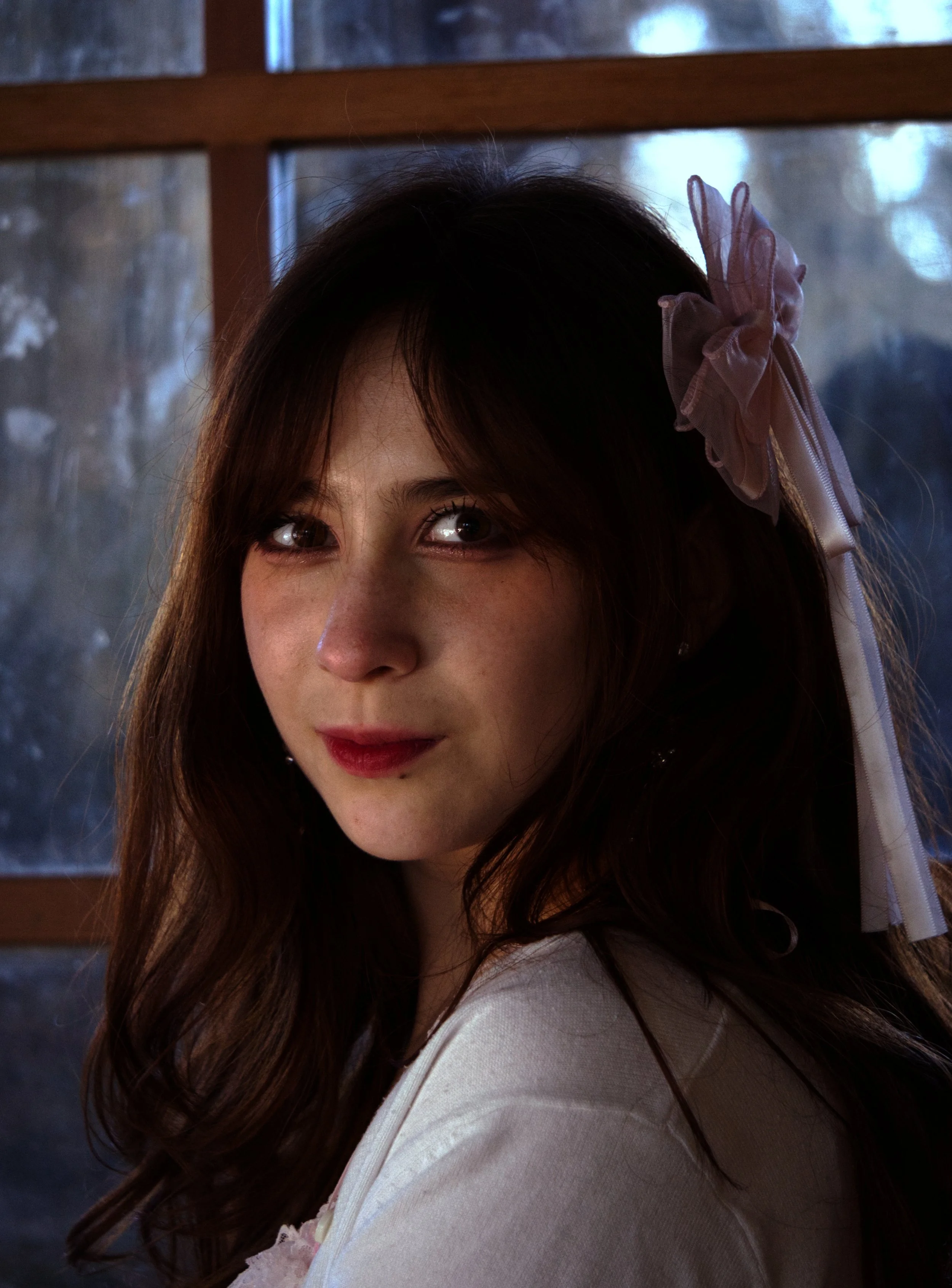 A young woman with long brown hair wearing a light pink or white top and a sheer bow in her hair, smiling gently with a rustic wooden window in the background.