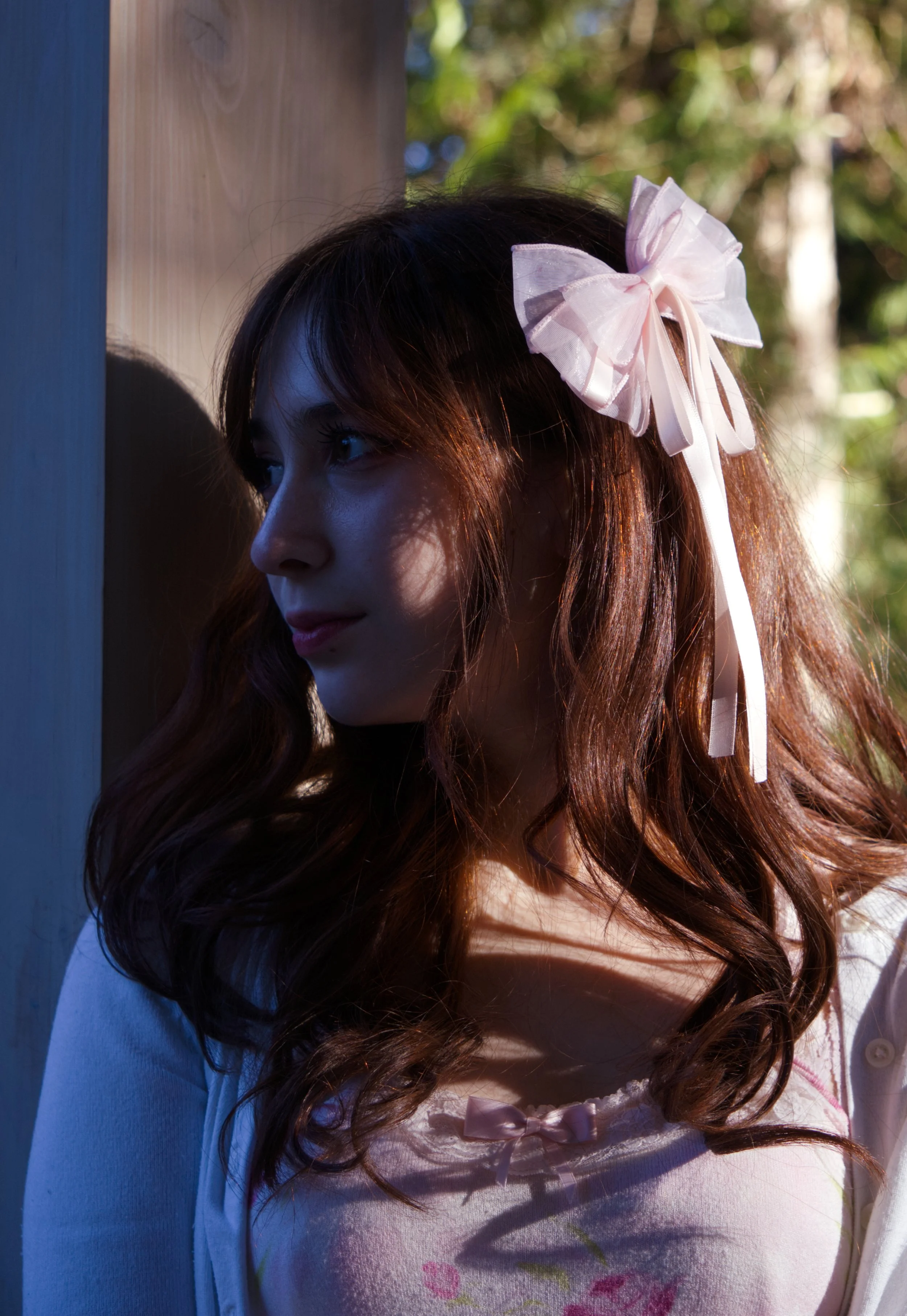 A young woman with long, wavy brown hair wearing a pink dress with a small bow and a white cardigan, standing outdoors with sunlight casting shadows on her face, a pink bow in her hair, next to a wooden post with a background of foliage and trees.