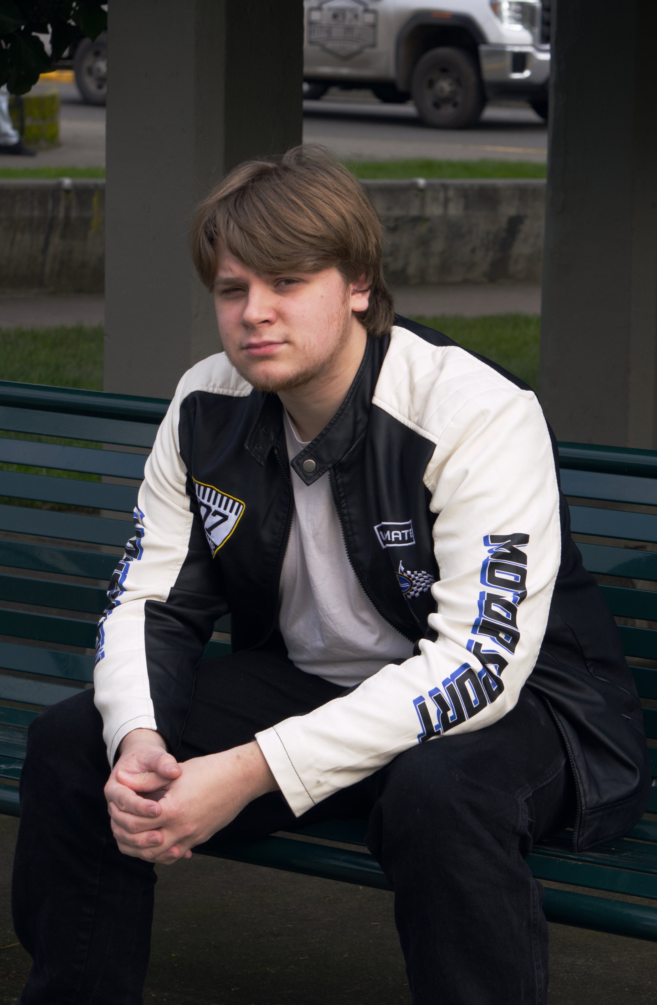 A young man with light brown hair, sitting on a park bench, wearing a black and cream jacket with racing patches, looking at the camera with a serious expression.
