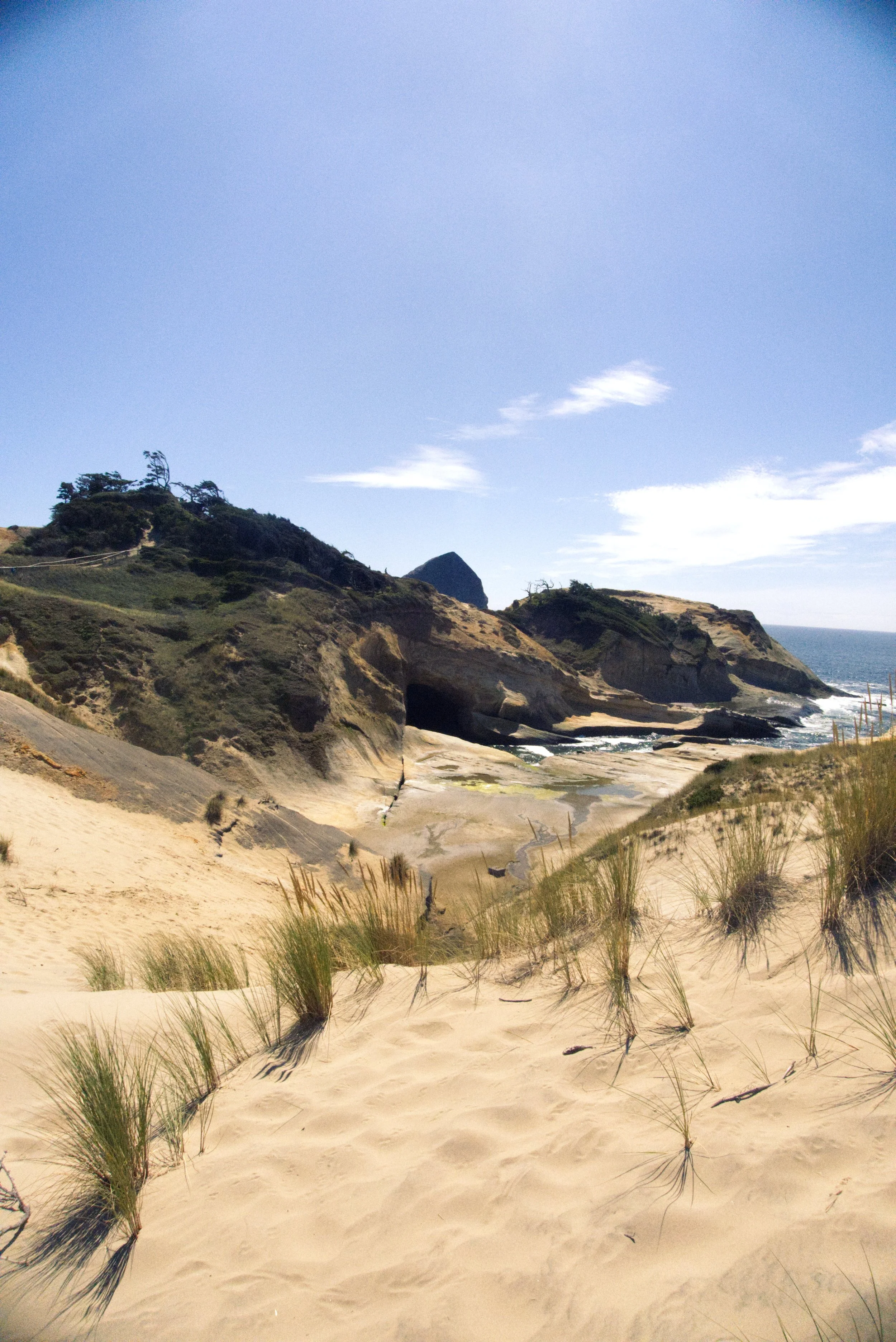 Sandy beach with dunes and grasses, rocky cliffs, and the ocean under a partly cloudy sky.