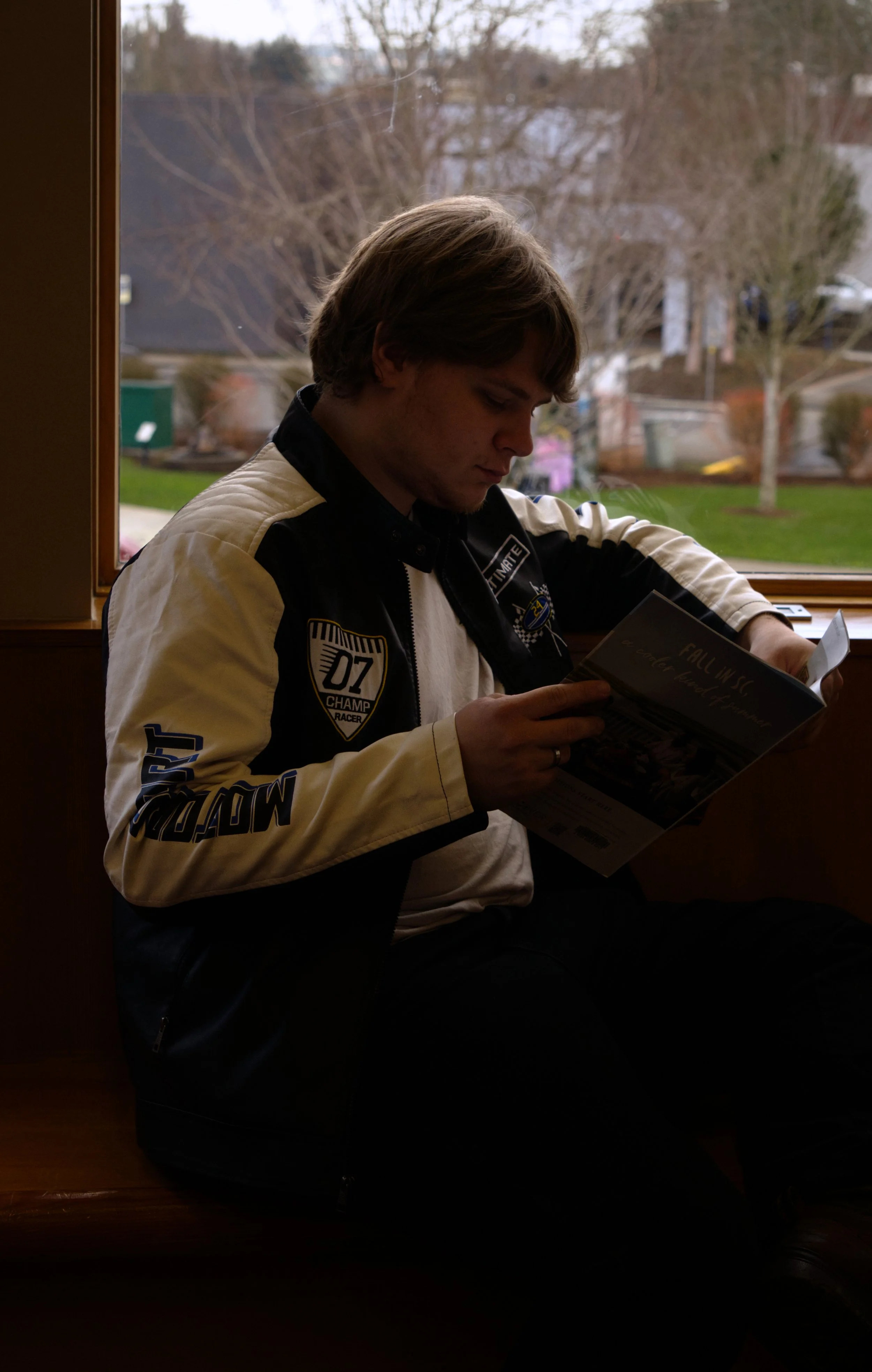 A young man sitting by a window, reading a brochure or magazine, wearing a black and white racing-style jacket with patches, against a background of leafless trees and buildings outside.