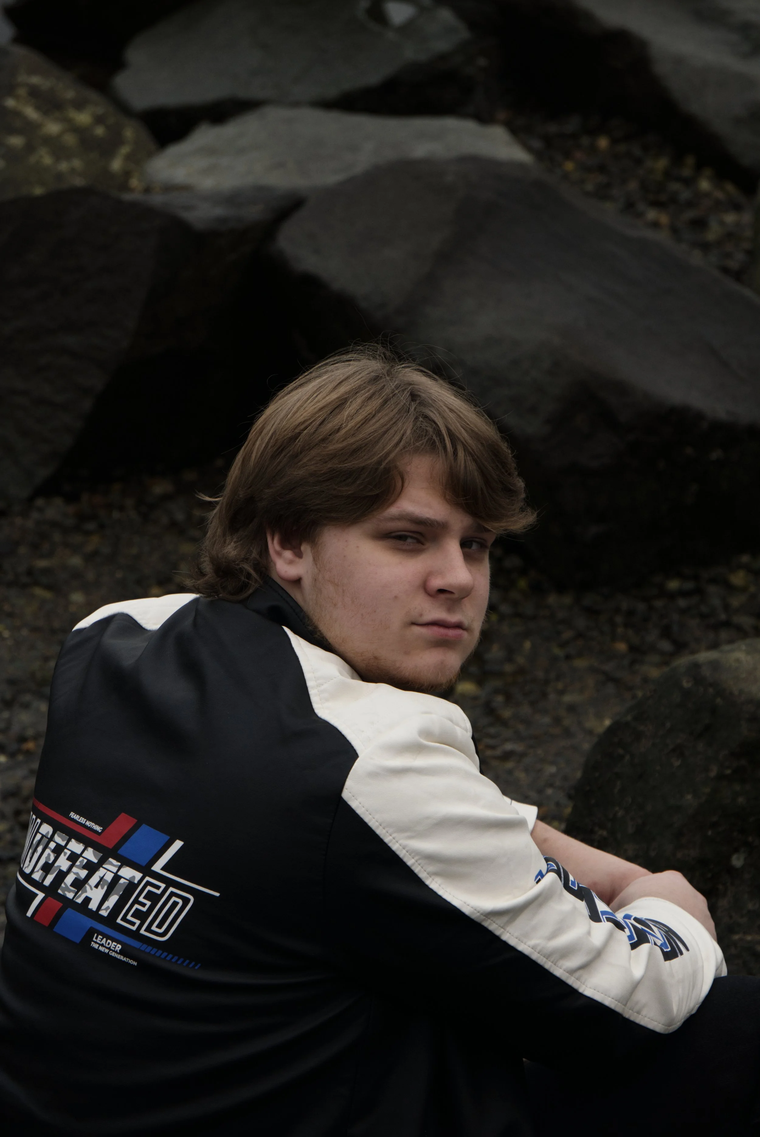 Young man with medium-length light brown hair sitting outdoors in front of large rocks, wearing a black and white jacket with red, blue, and white text and graphics.