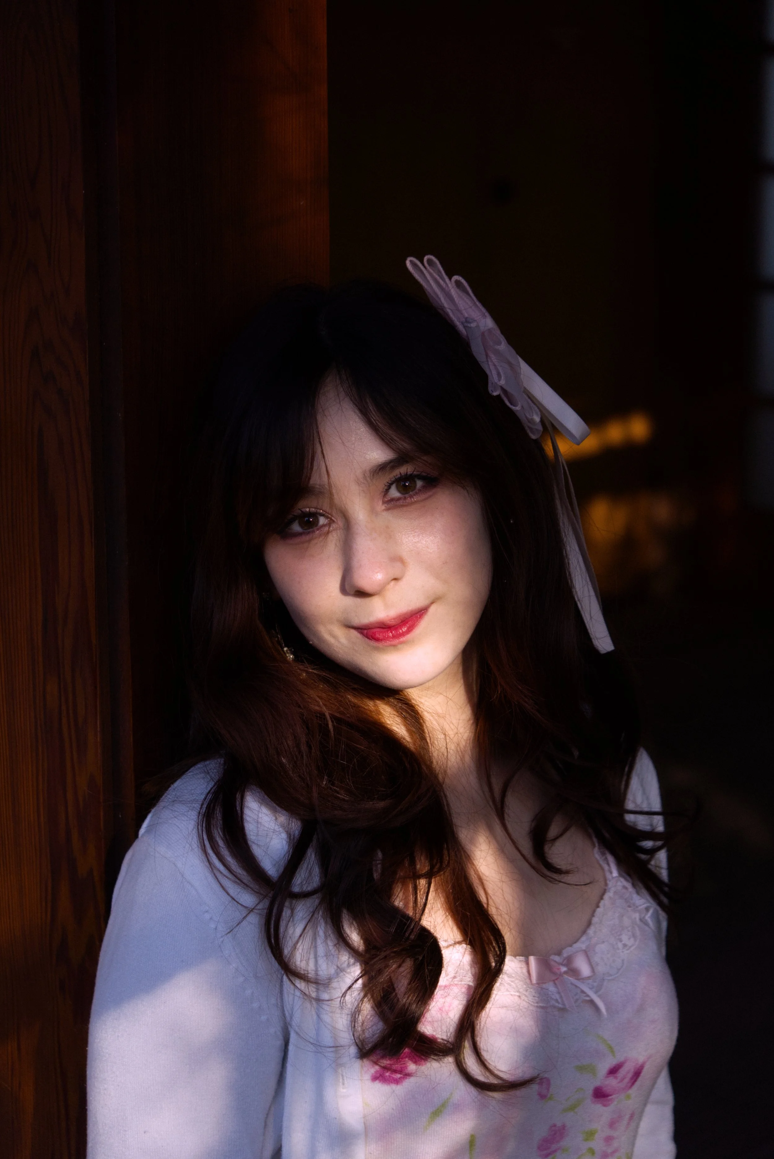 A young woman with dark hair, light makeup, and a pink bow on her dress, posing against a wooden wall in a dimly lit environment.