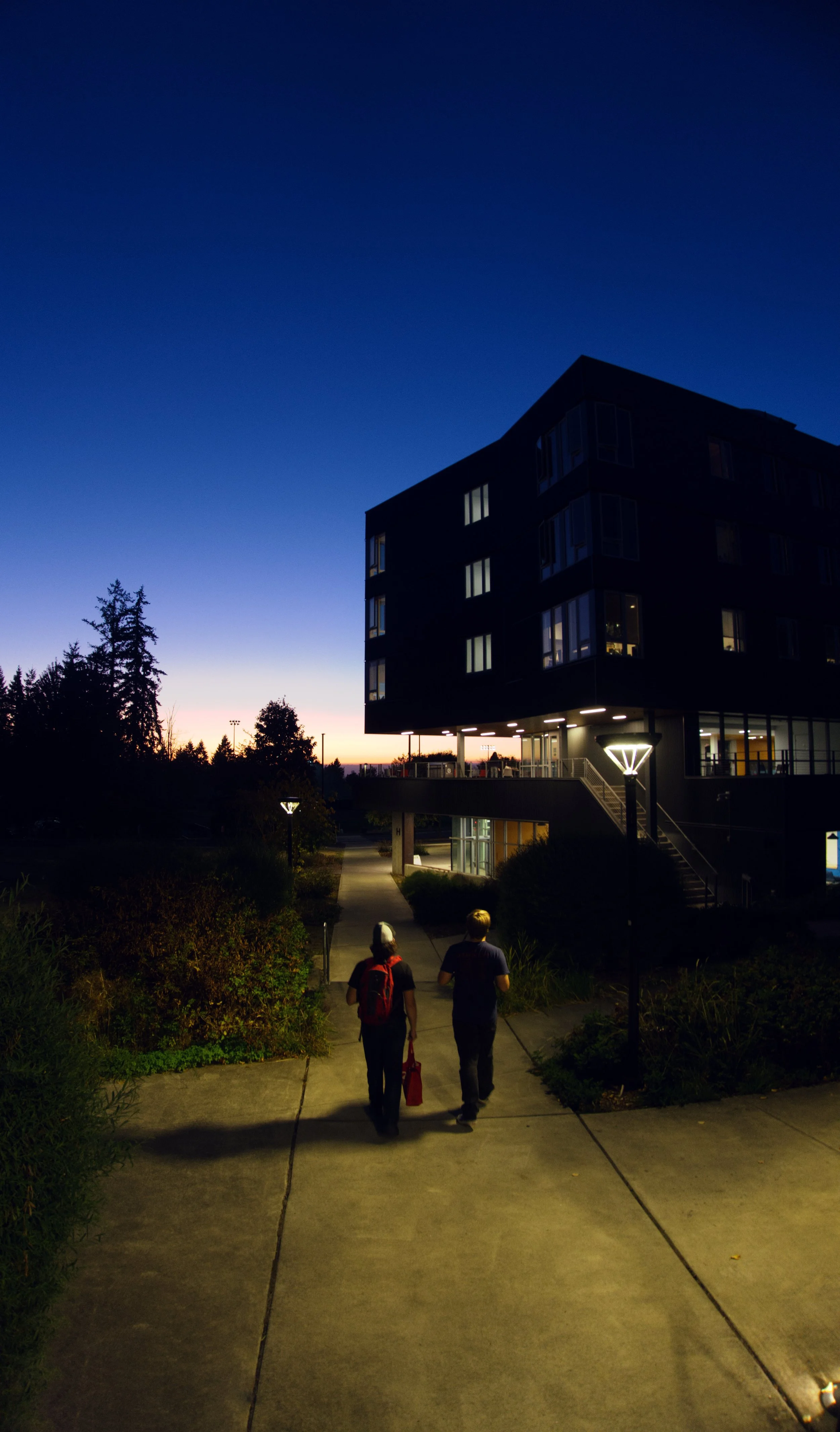 Two people walking on a sidewalk in front of a modern apartment building at dusk. The sky is transitioning from sunset to night, and the area is illuminated by streetlights.