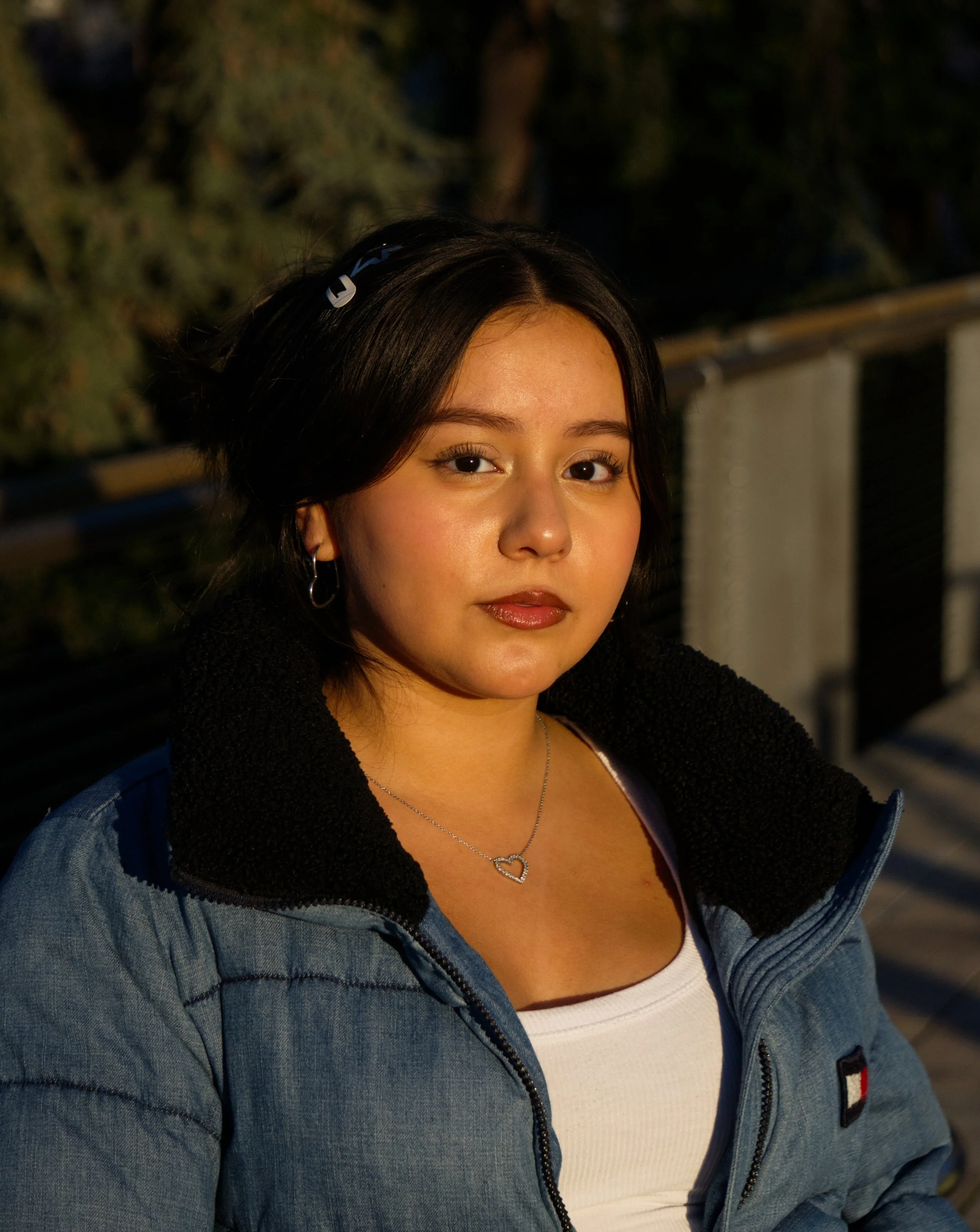 A young woman with dark hair, wearing a denim jacket over a white top, silver jewelry, and hoop earrings, sitting outdoors during sunset.