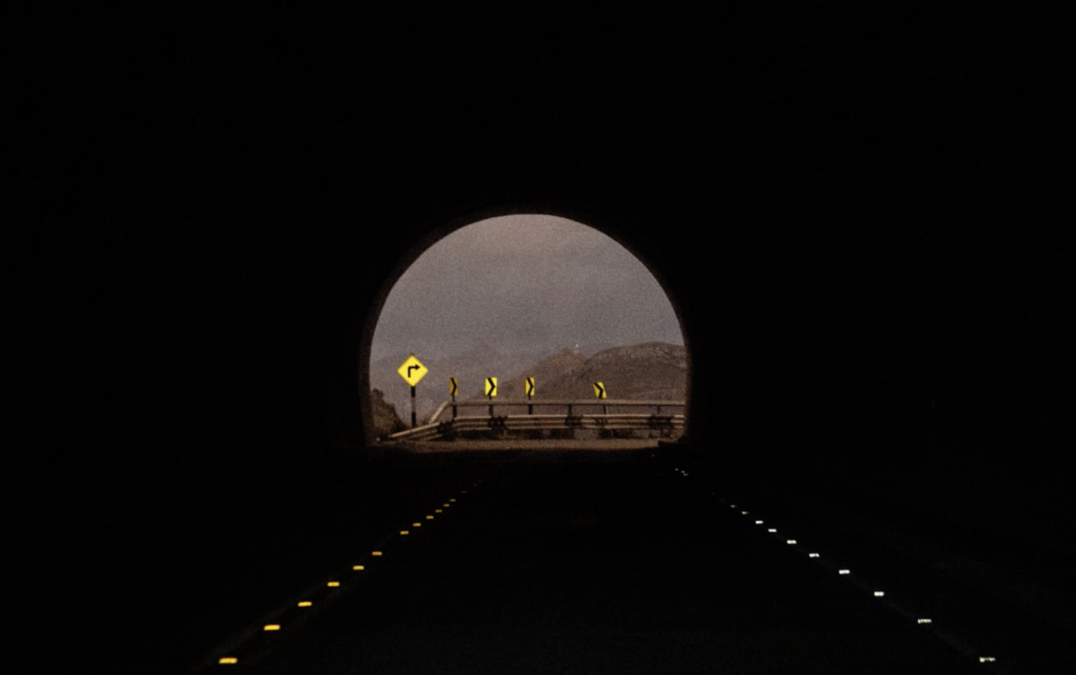 View through a dark tunnel showing a winding mountain road outside with yellow and black directional signs.