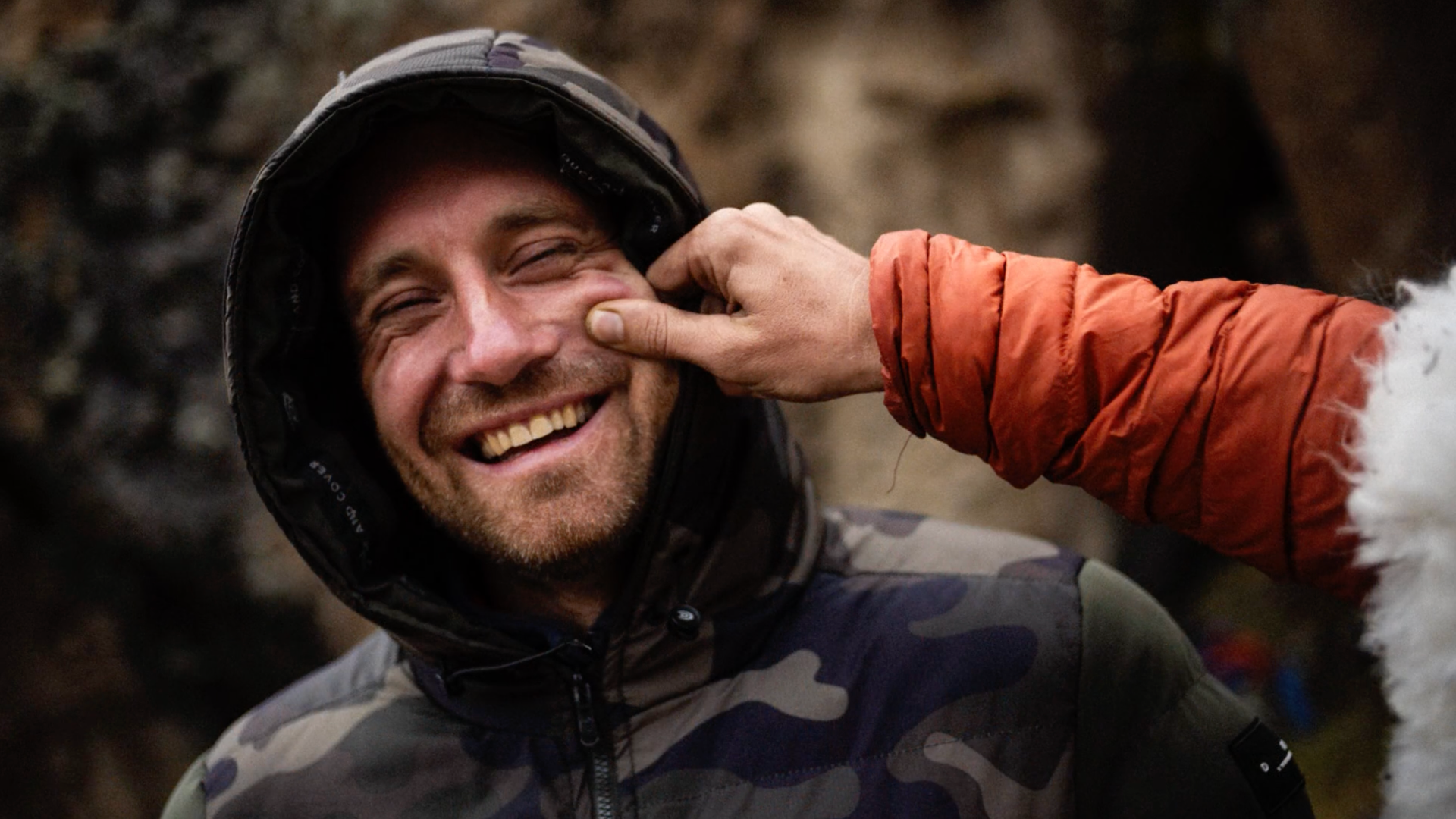 A man is smiling with a camouflage jacket and hood, while a second person, wearing an orange jacket, is pinching his cheek with a finger.