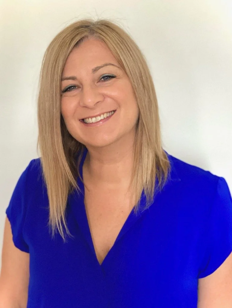 A woman with blonde hair smiling, wearing a royal blue top, standing against a plain light-colored background.