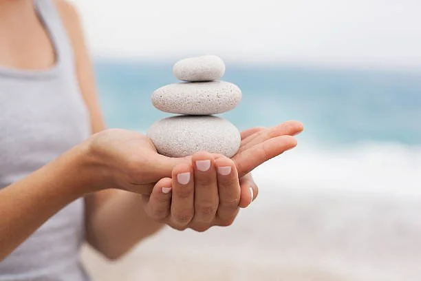 Person holding four stacked smooth stones on a beach with ocean in the background.