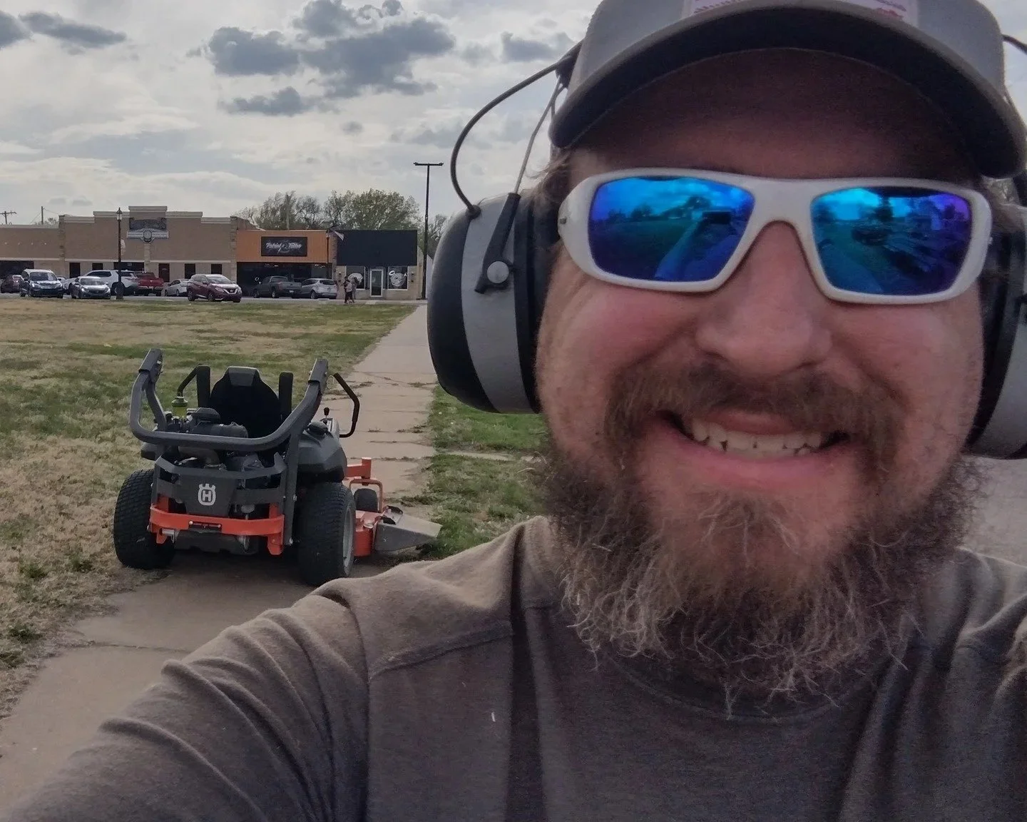 Close-up of a smiling man with a beard, wearing sunglasses, a cap, and earmuffs, taking a selfie outdoors with a lawn mower and strip mall in the background.
