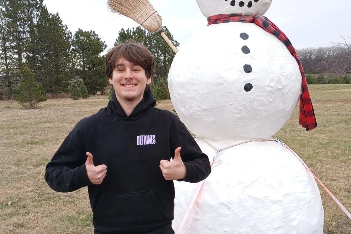 A young man standing outdoors next to a large snowman, giving two thumbs up and smiling.