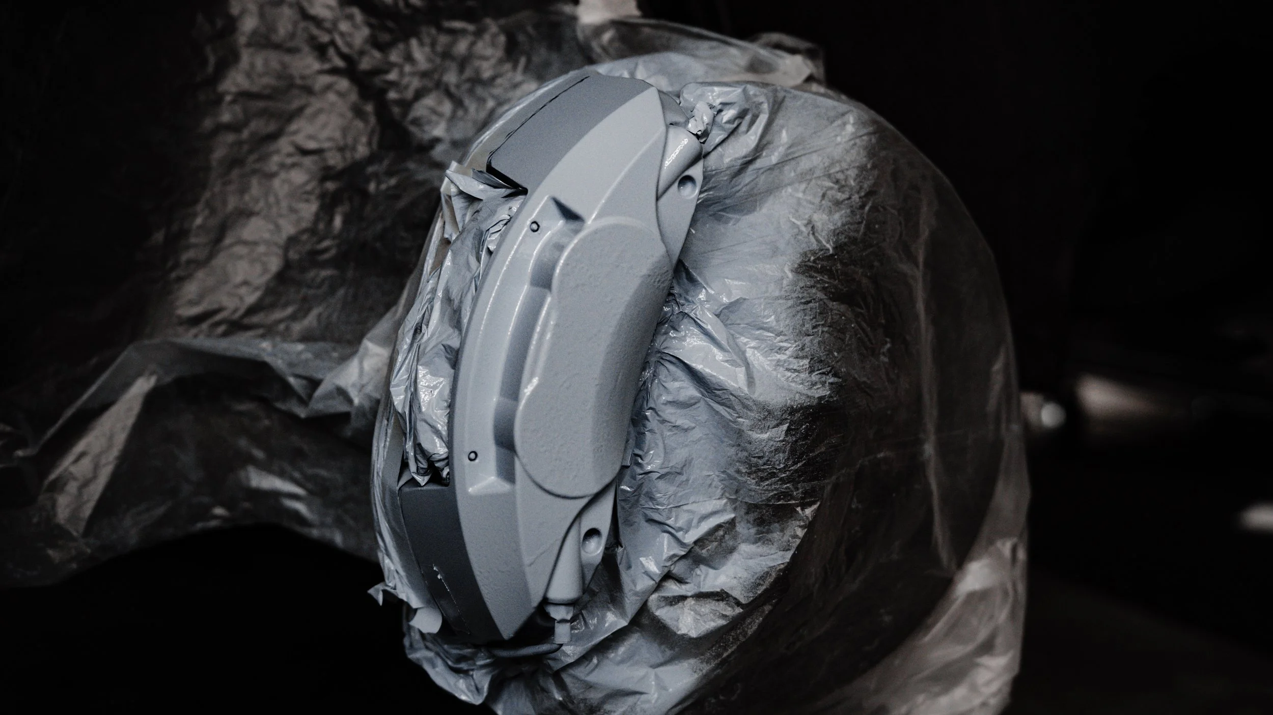 A partially unwrapped gray plastic object, possibly a device or machinery part, on crumpled black foil or plastic in a dark environment.