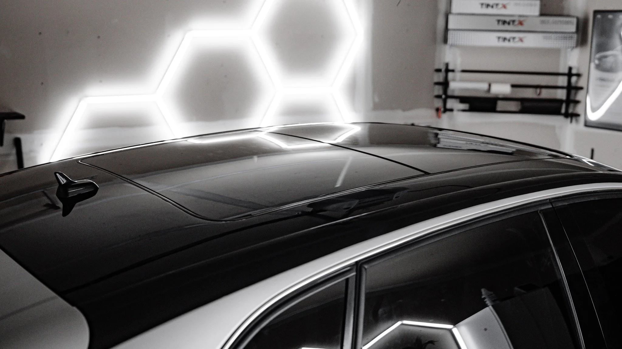 A black car with a sunroof parked inside a garage with honeycomb-patterned LED wall lighting.