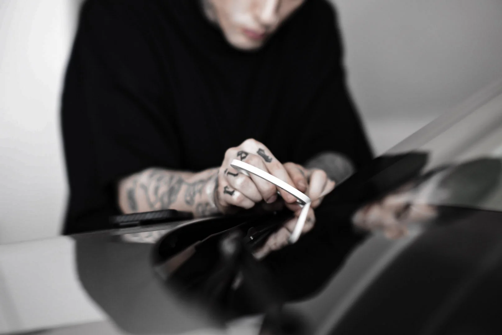 A person with tattoos on their arms is using a white smartphone on a reflective black surface.