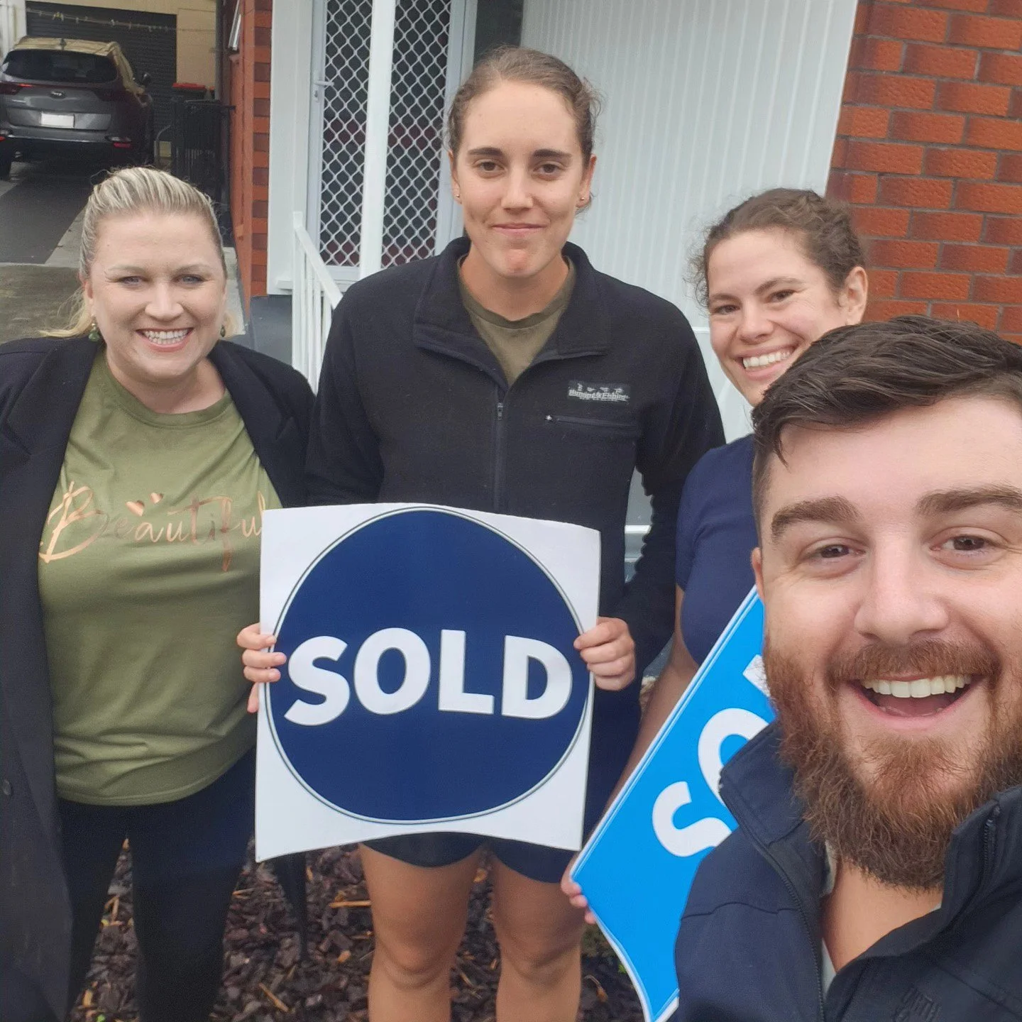 Four people standing outside, smiling and holding signs that read 'SOLD'.