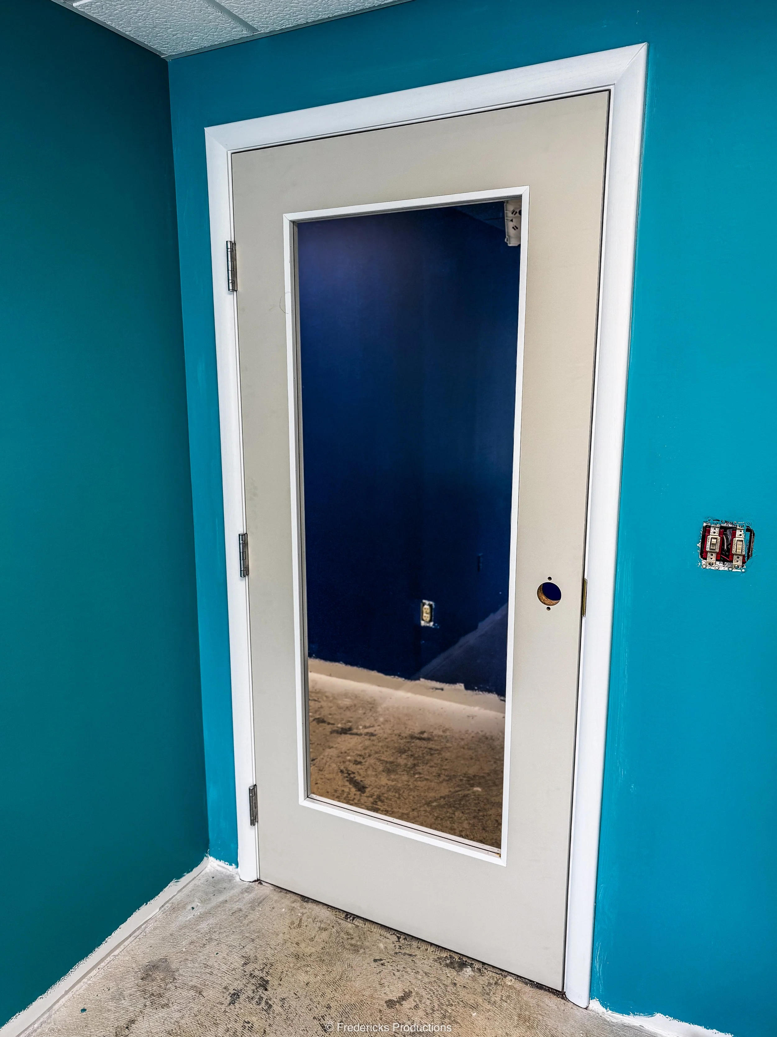A white door with a glass panel in the center, leading to a room with dark blue walls. The door is slightly ajar, and there are electrical outlets and a switch box on the blue wall adjacent to the door. The floor appears to be under construction or r