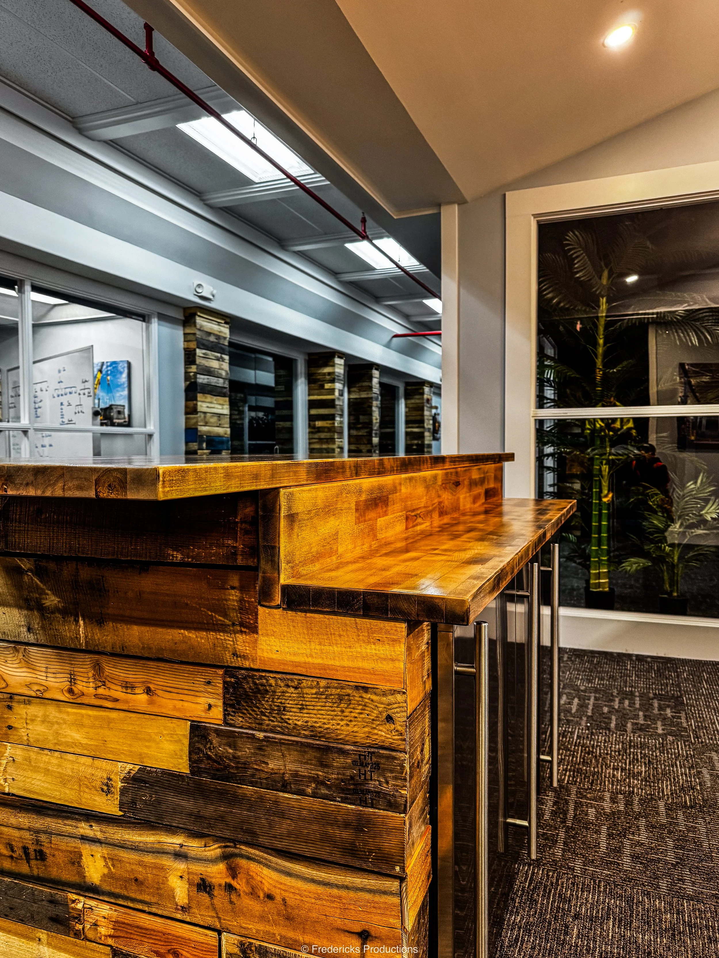 Interior of a modern office or cafe with a wooden free-standing counter, large windows, and potted plants outside the window.