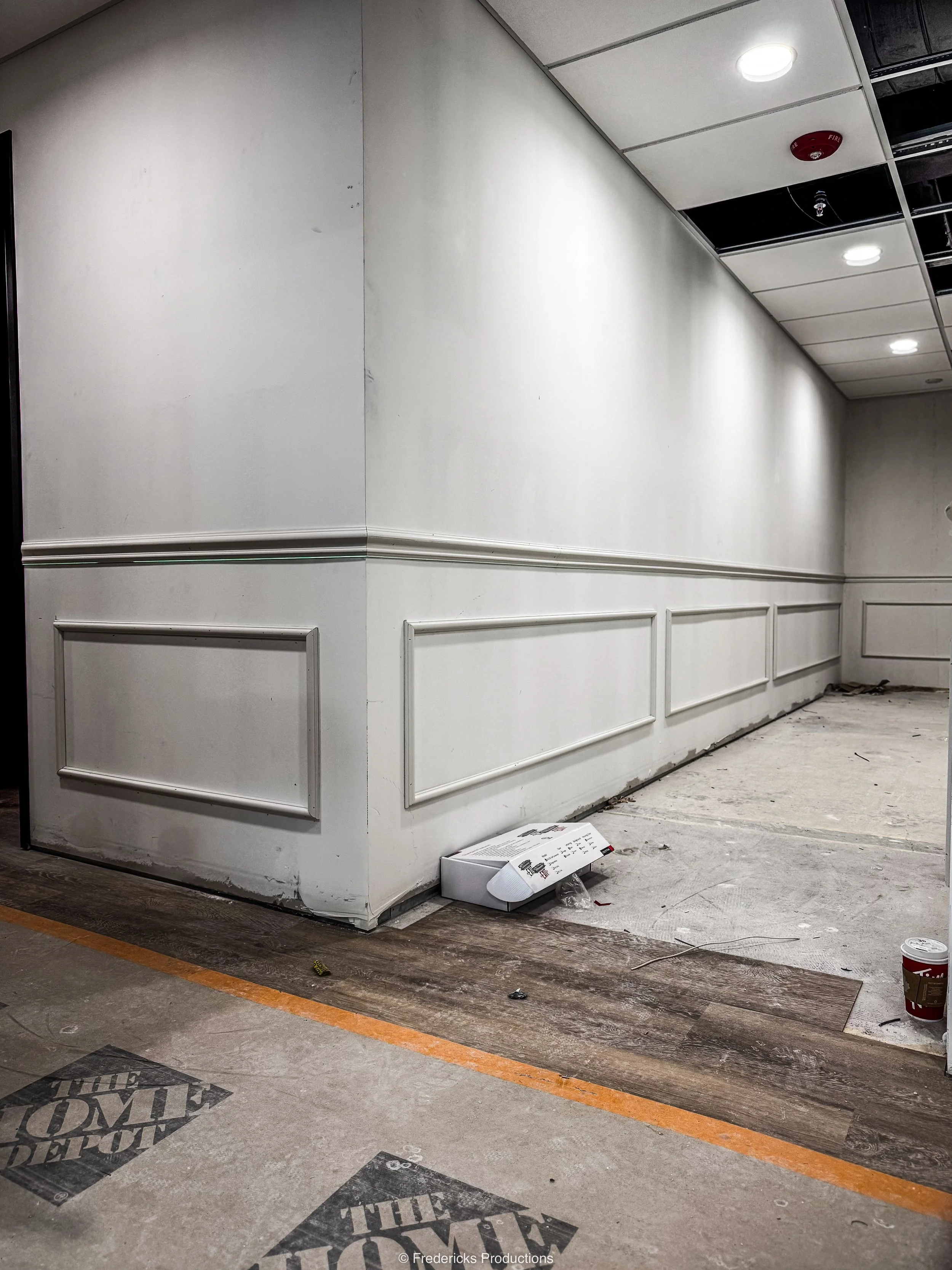 Empty interior space with partially removed flooring, white wall paneling, construction debris, and a coffee cup on the floor.