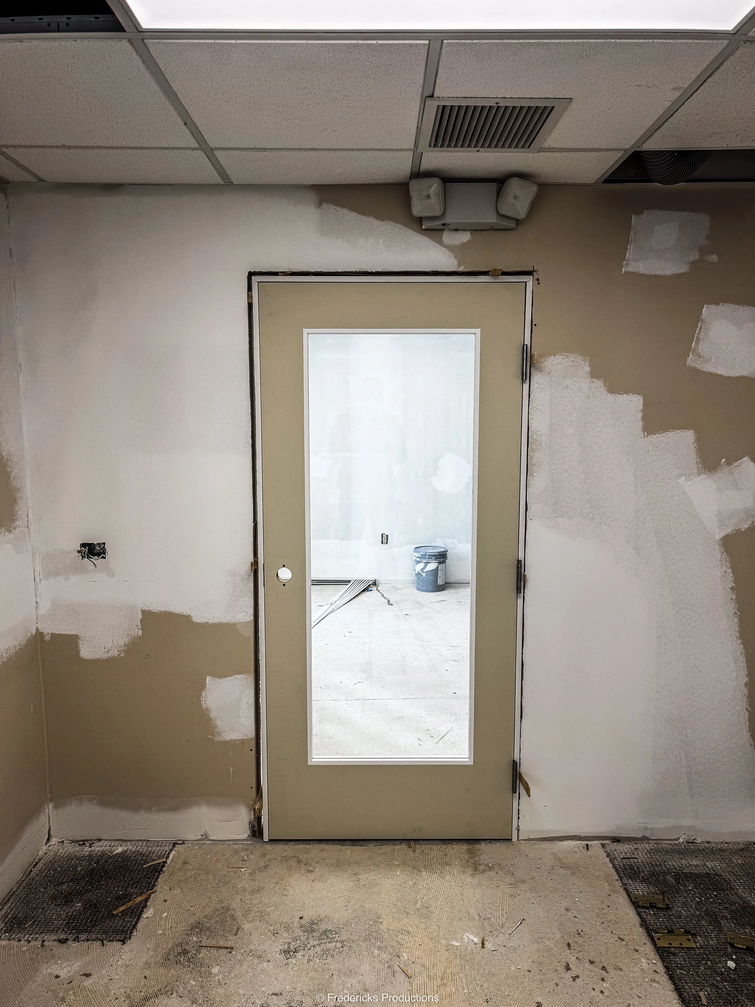 Interior wall under construction with drywall patching, a door with a large glass window, ceiling tiles, and construction materials on the floor.