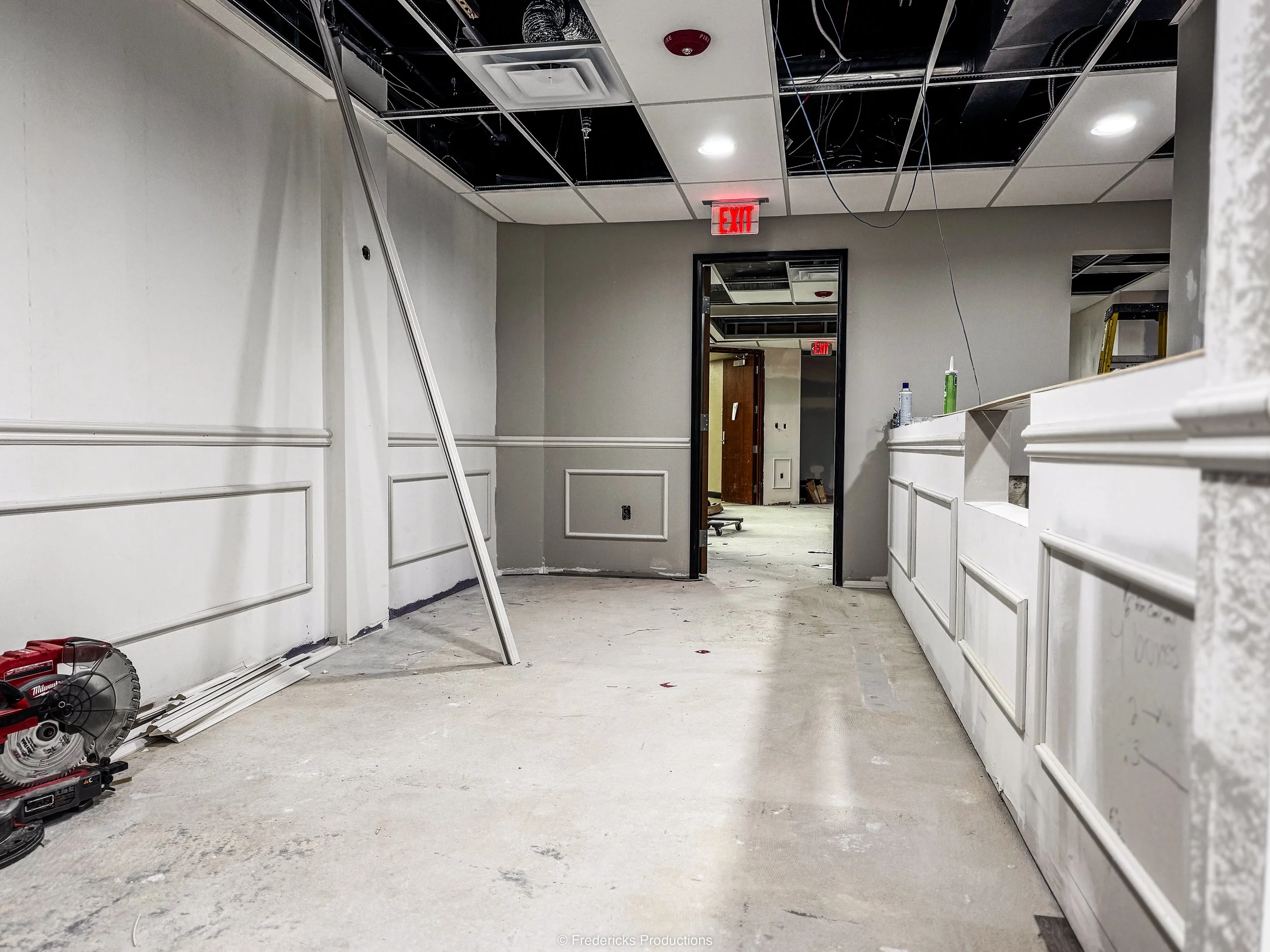 Interior of a room under renovation with construction tools, partially finished walls, an open doorway, and exposed ceiling panels and wiring.