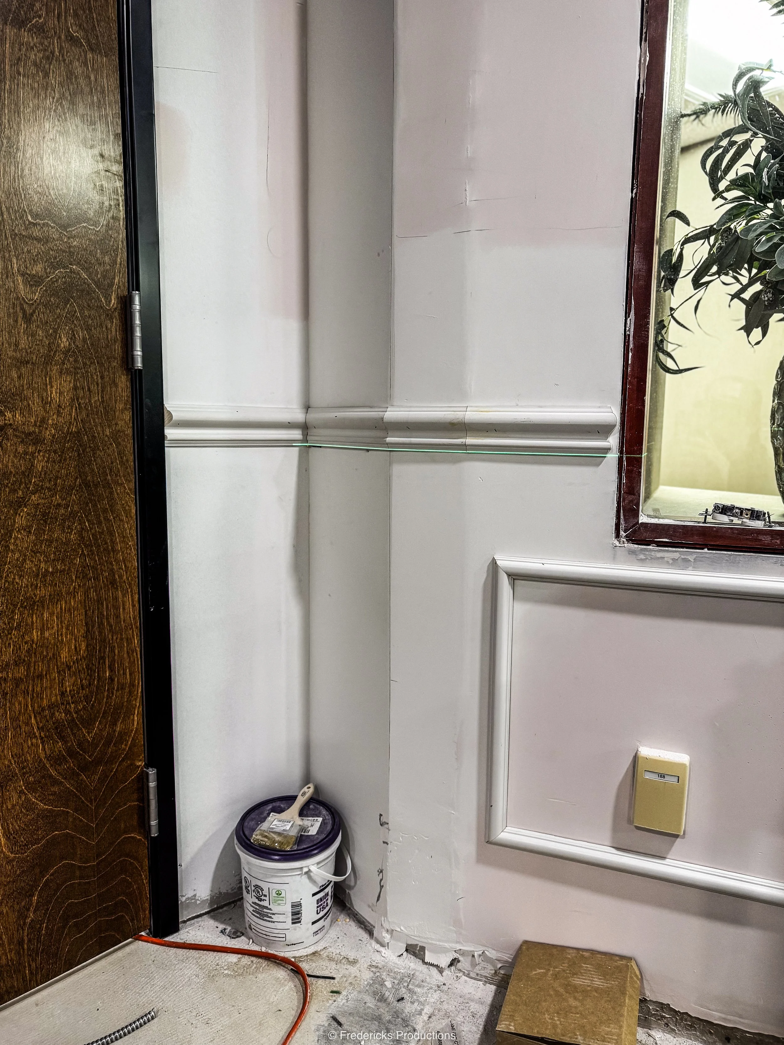 Corner of a room with a wall, window, and door, showing construction or renovation work, with a bucket, some tools, and an orange cord on the floor.