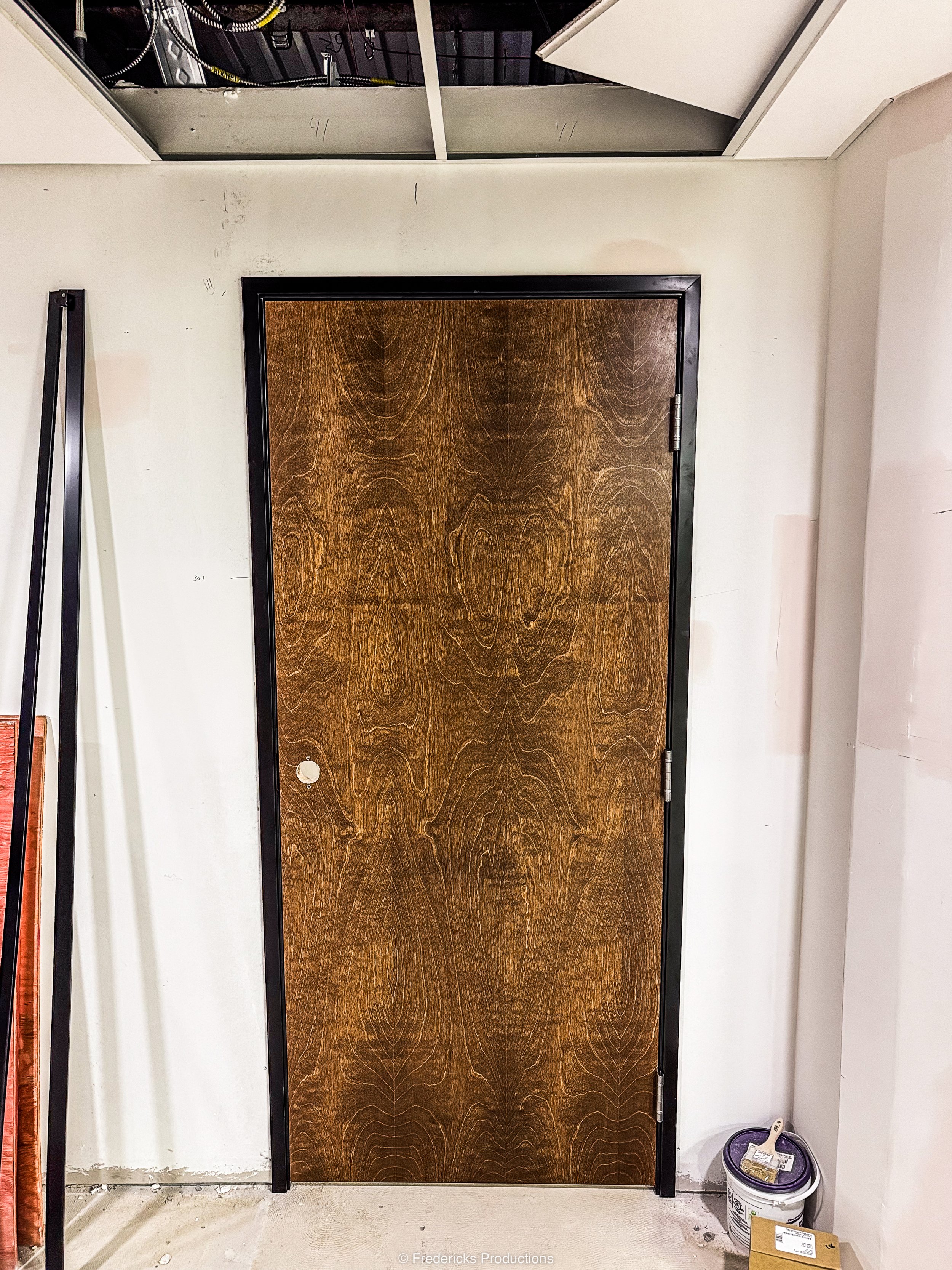 A closed wooden door with a dark frame in a room under construction, with a bucket and paintbrush on the floor nearby.