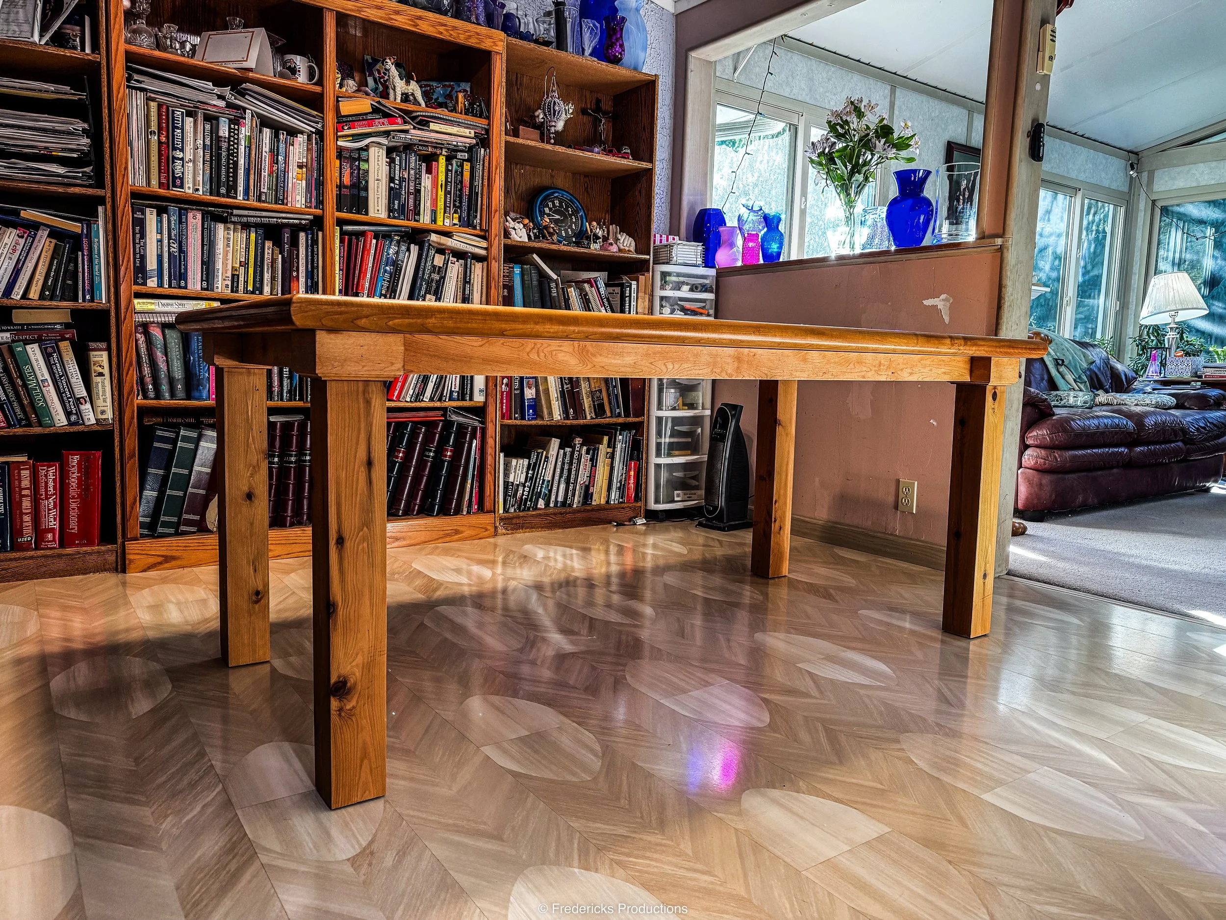 A wooden dining table in a room with a bookshelf filled with books and decorative items, next to a hallway or living room with large windows, blue and purple vases, and a couch with a blanket and cushions.
