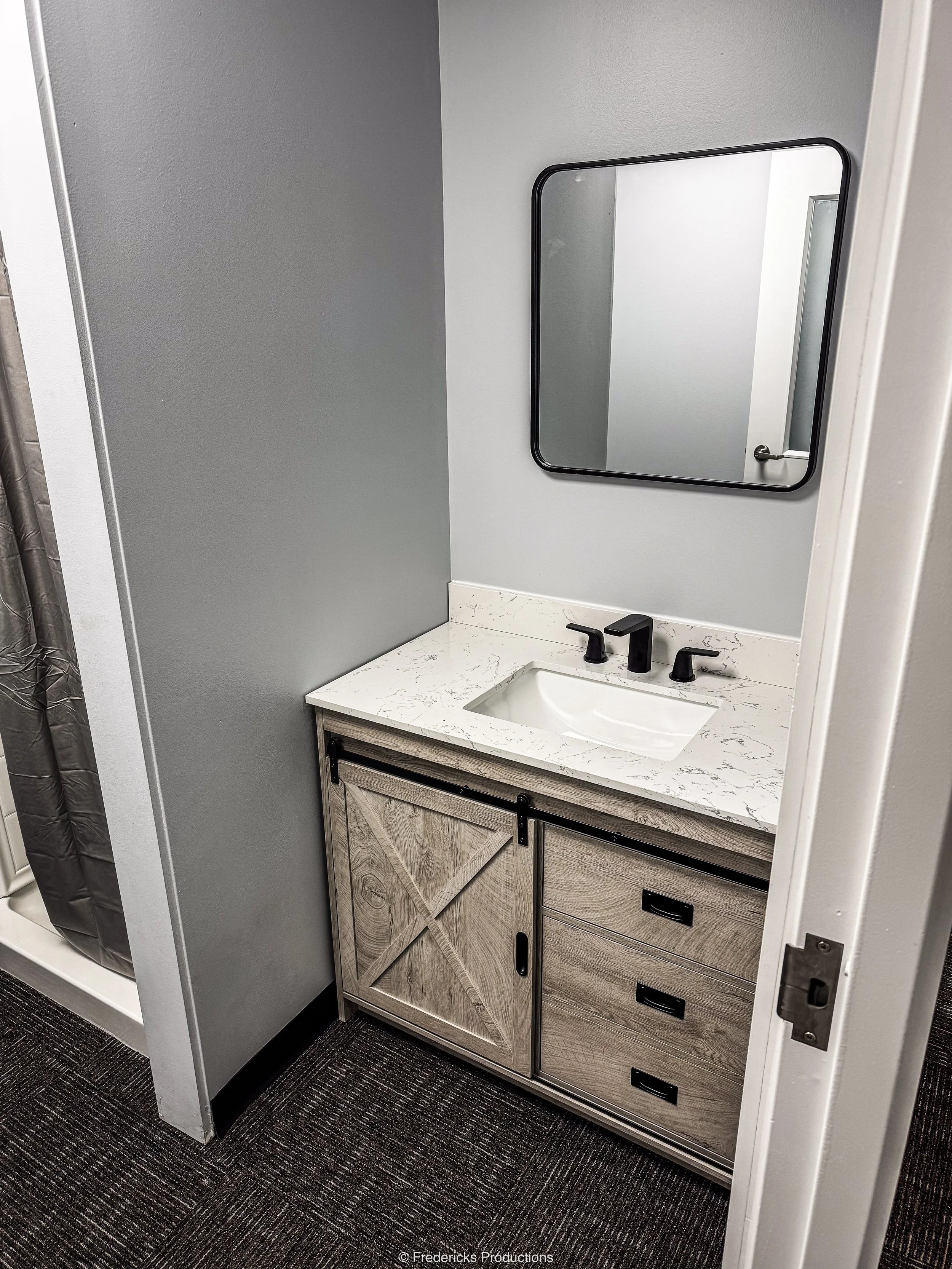 Small bathroom vanity with a rectangular sink, black faucet, wood cabinet with sliding barn door and three drawers, wall-mounted mirror, gray wall, and dark carpet.