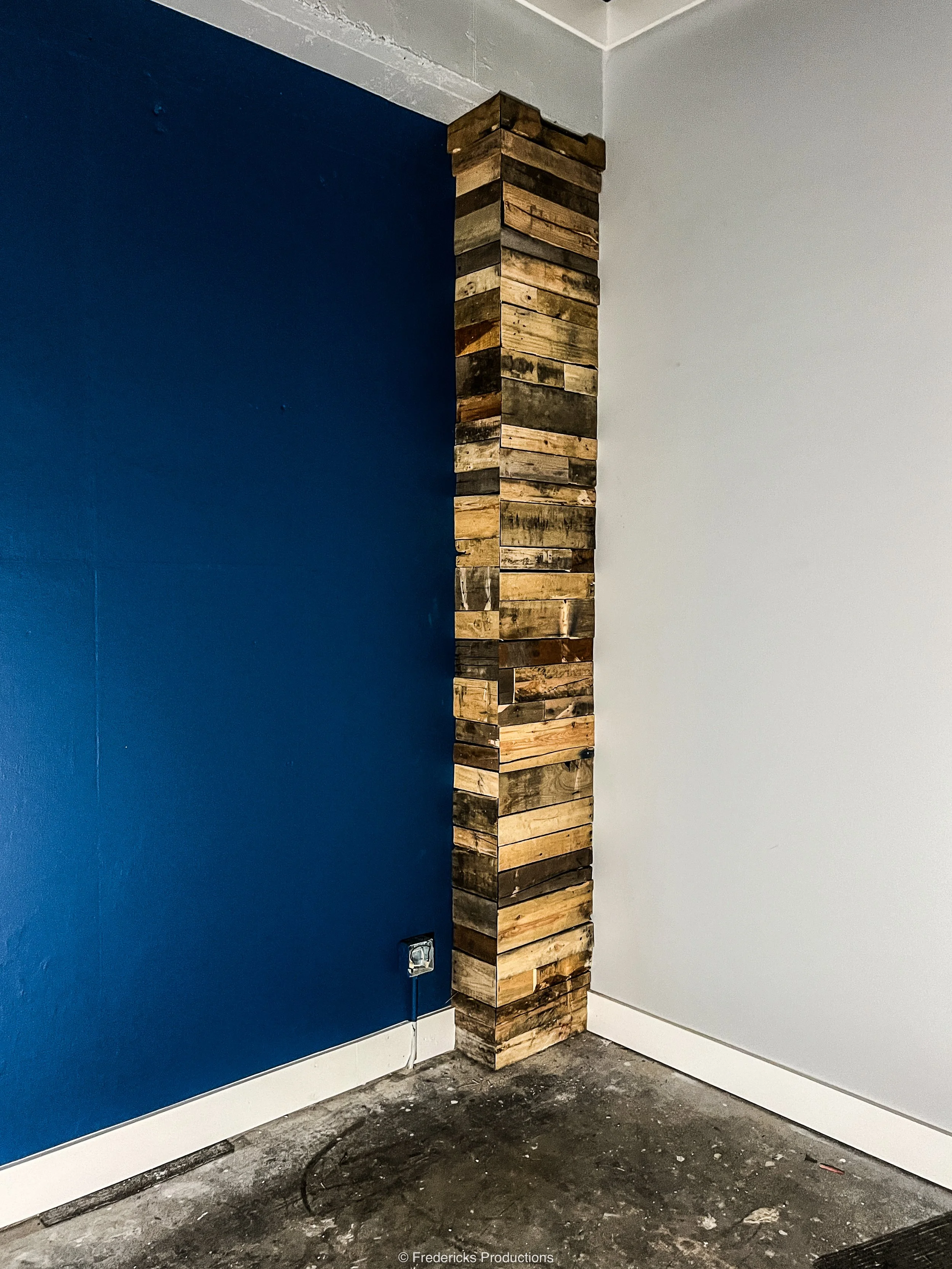 Corner of a room with a blue wall on the left, a white wall on the right, and a wooden pallet pillar in the corner. There is an electrical outlet at the base of the blue wall and a dark concrete floor.