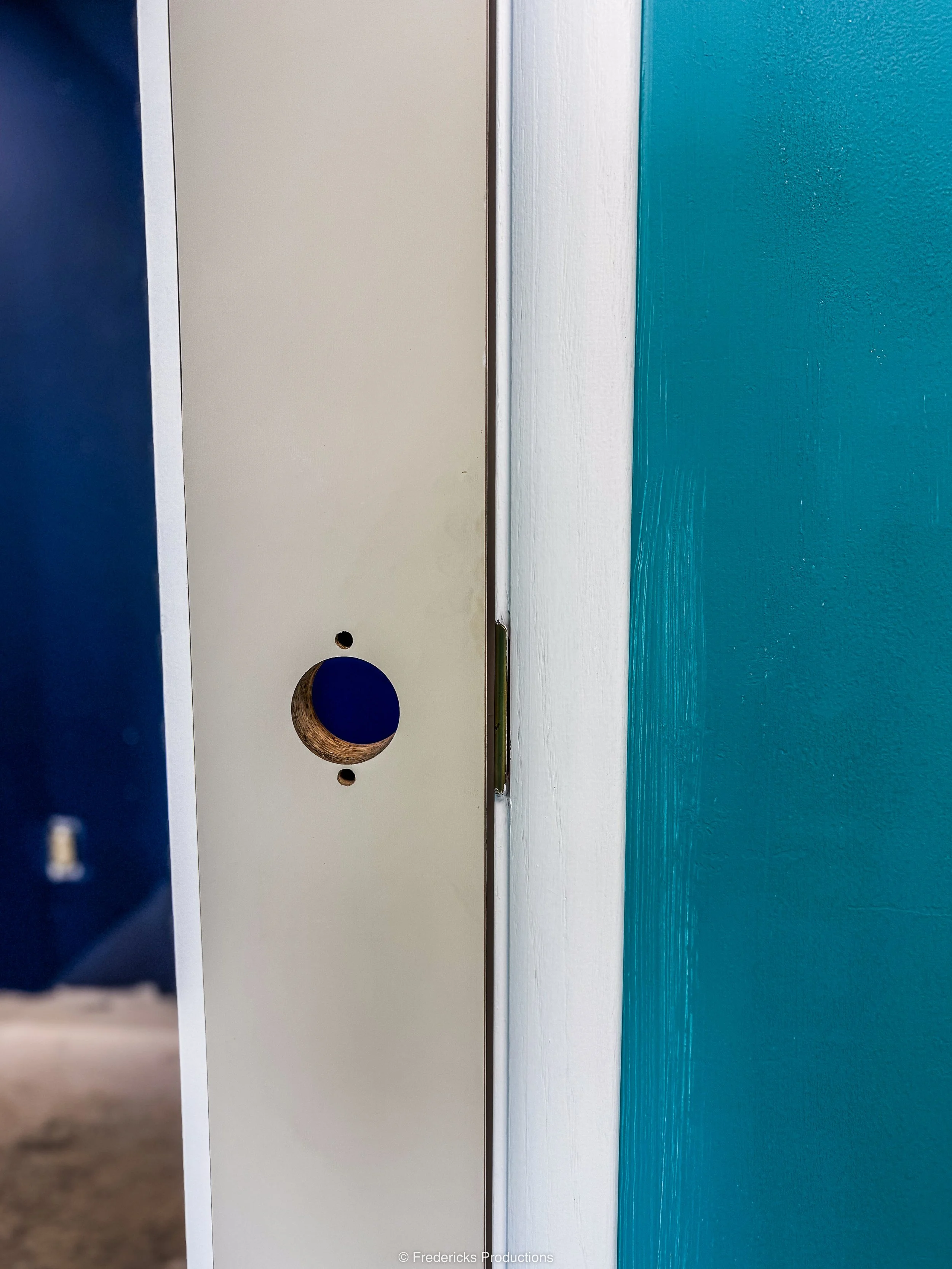 Close-up of a white doorframe with a hole in the door, showing a dark interior behind. The door is partially open, revealing a blue wall in the background.
