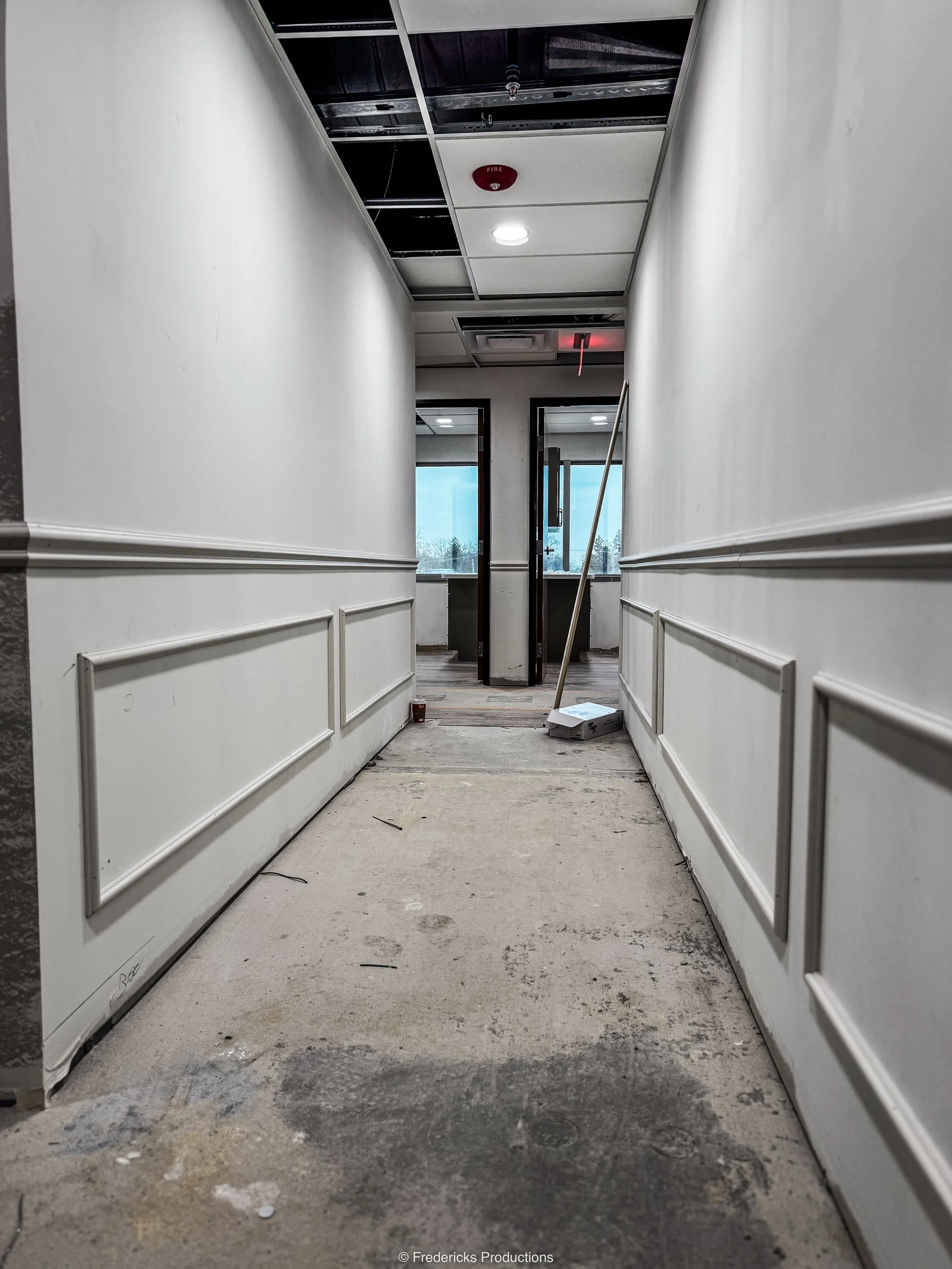 Interior hallway under renovation with partially finished ceiling, messy floor, and construction tools leaning against the wall.