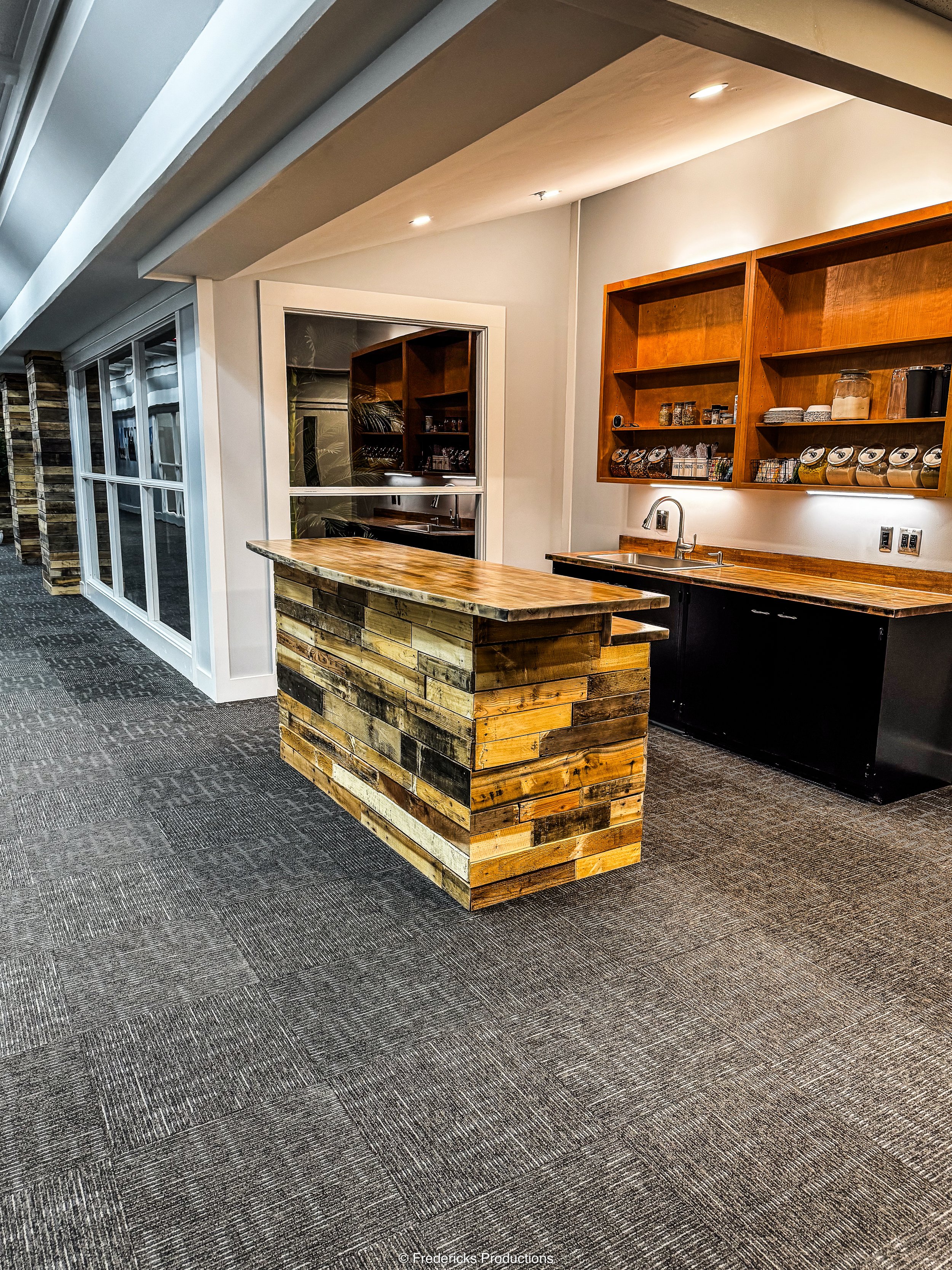 Interior of a modern kitchen or coffee bar area with a wooden counter made of reclaimed wood and open wall-mounted wooden shelves with jars and cups, a sink under the shelves, and a glass wall with sliding doors to the left.