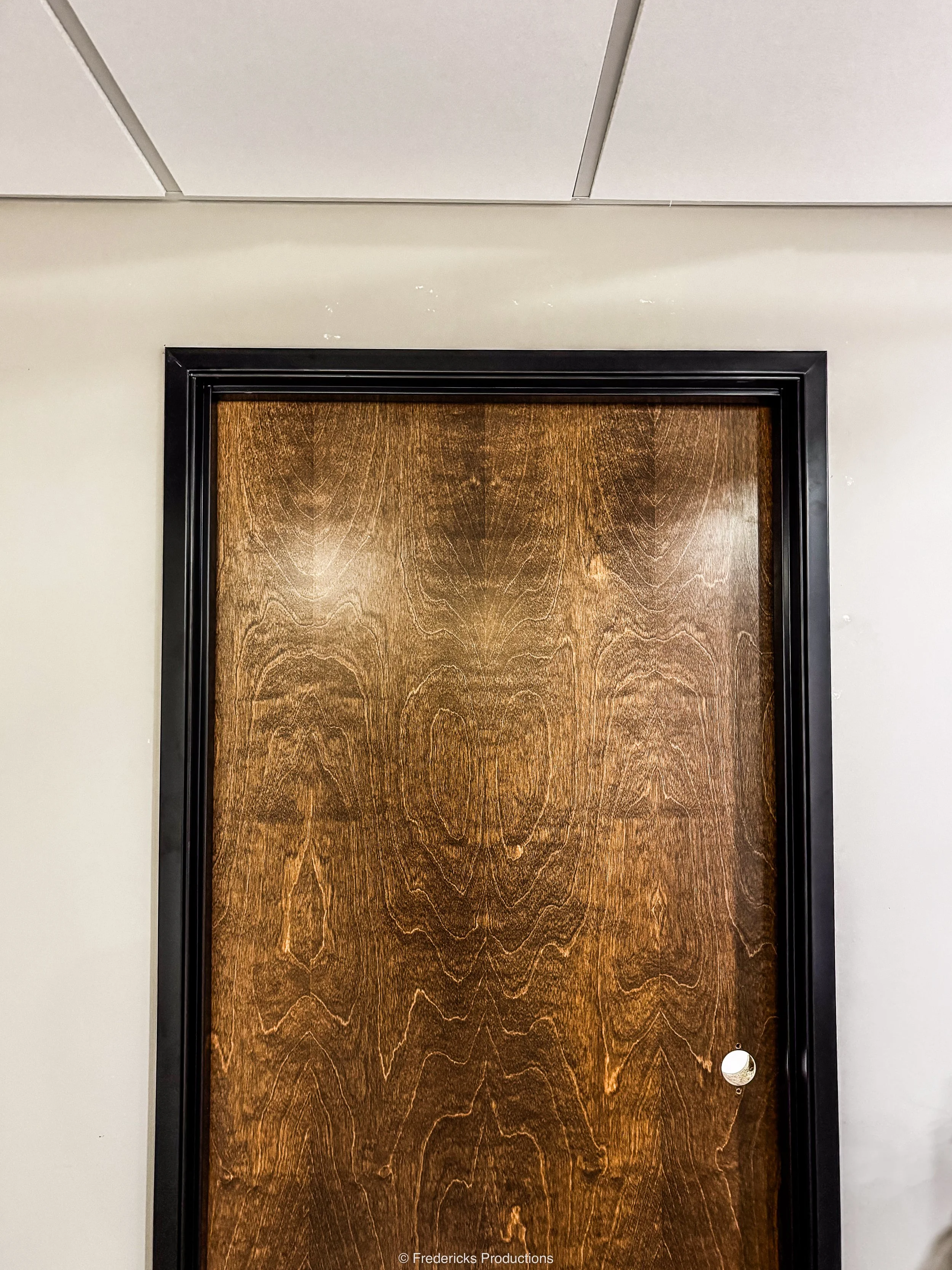 A wooden door with a black frame, in a room with a white wall and ceiling tiles.